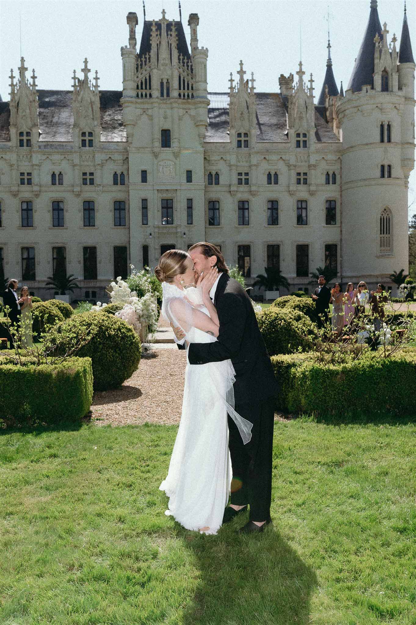 First kiss at Gothic chateau with bride cupping groom's face, white peony aisle arrangements on gravel