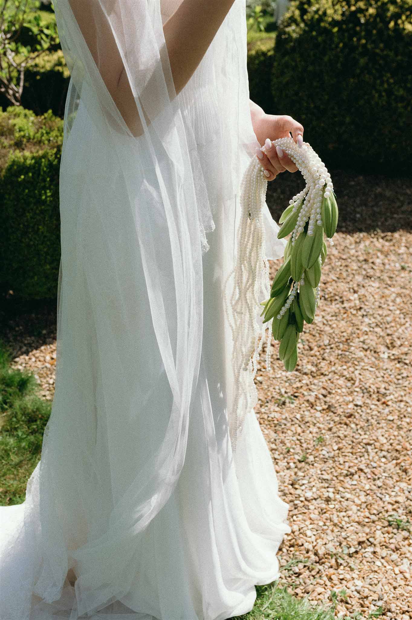 Bride from behind in open-back chiffon gown holding cascading pearl strand bouquet with olive leaves