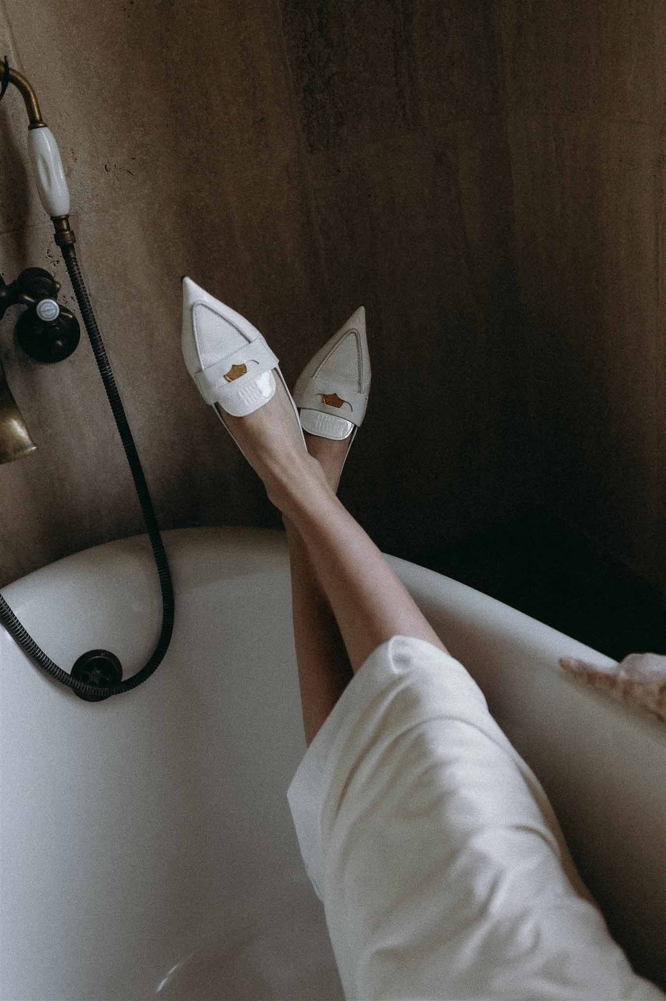 Bride's white patent loafers with gold crown emblem propped on clawfoot bathtub rim in moody light