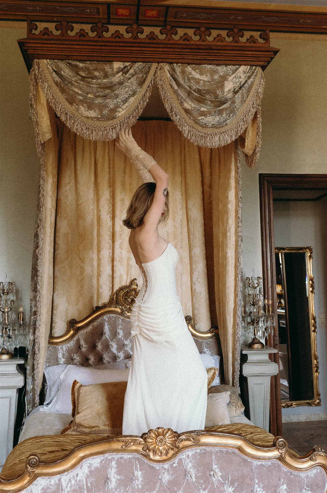 Bride in strapless ivory gown reaching toward gold canopy of ornate four-poster bed in chateau bedroom