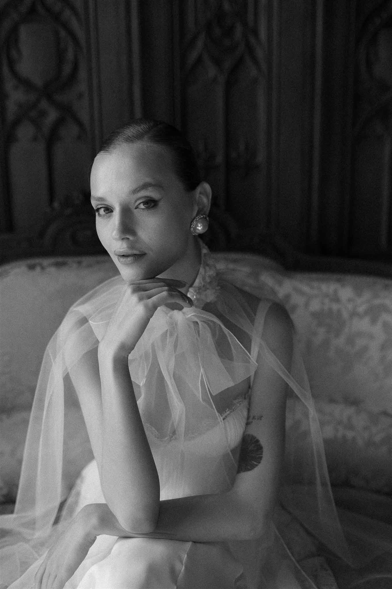 Black and white portrait of bride in sheer tulle cape with pearl cluster earring on settee