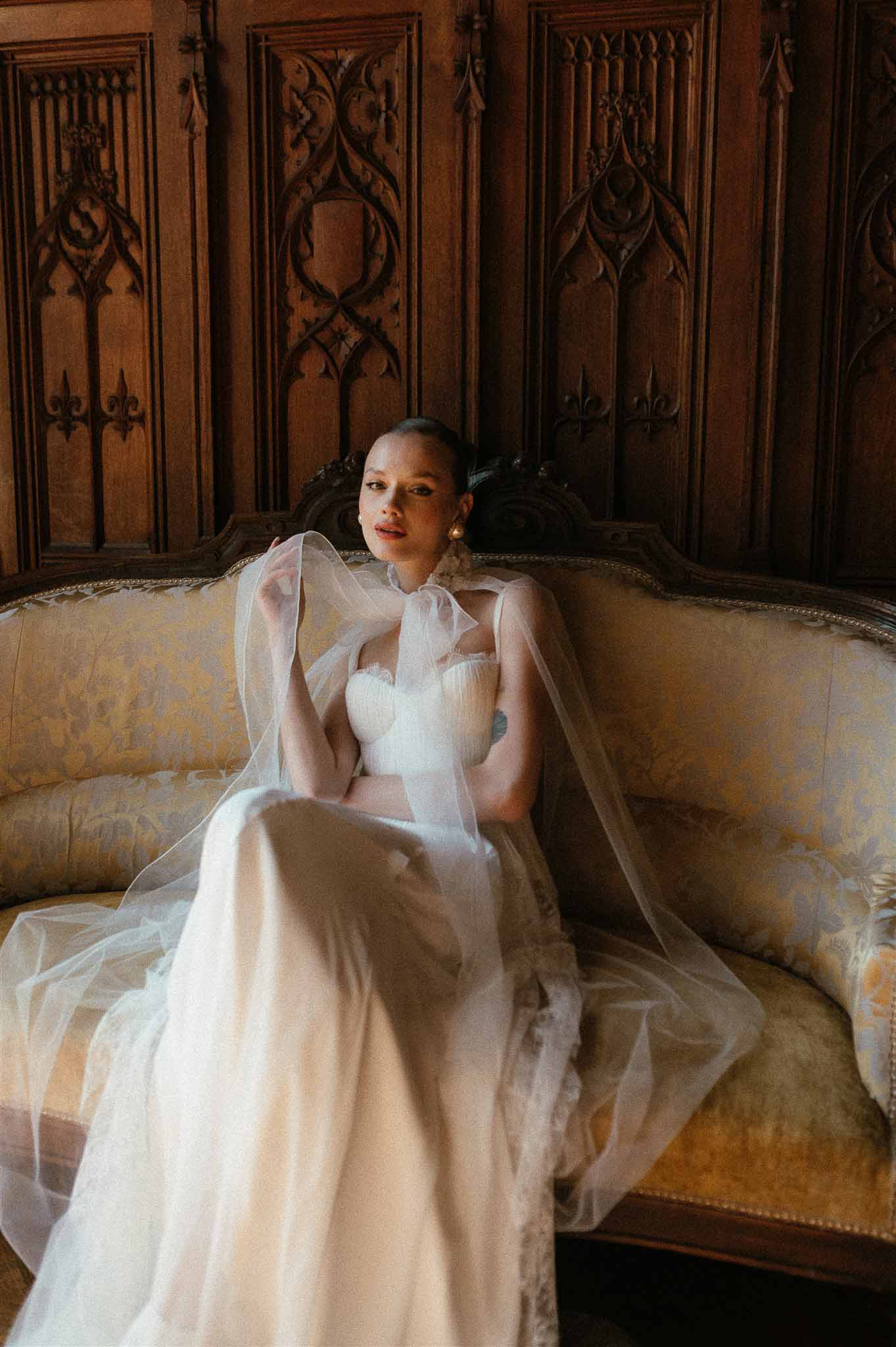 Bride in ivory corset gown with sheer tulle cape on gold damask sofa in Gothic carved wood interior