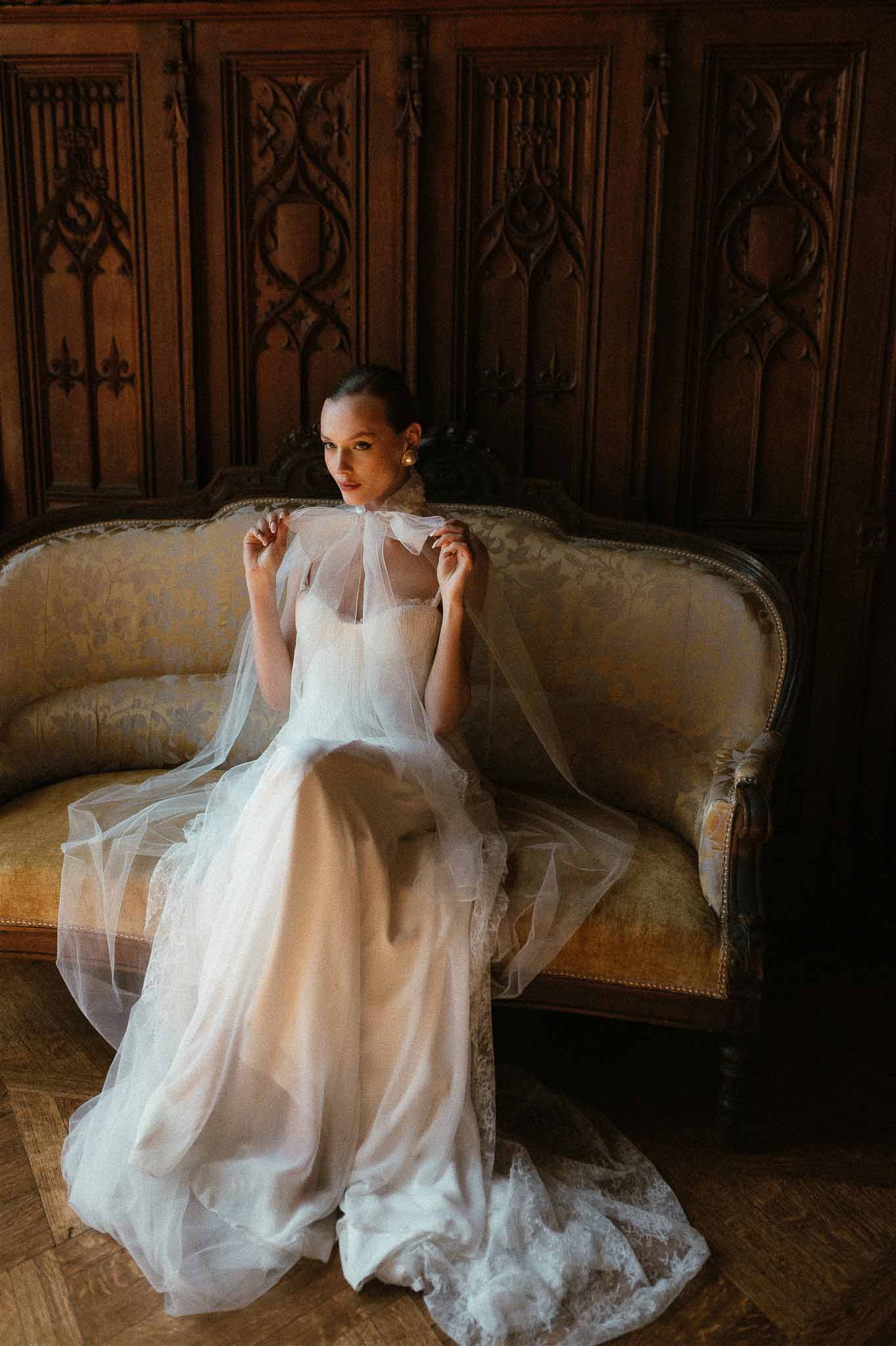 Bride in tulle gown with sheer bow-tie cape on gold damask loveseat before Gothic carved paneling