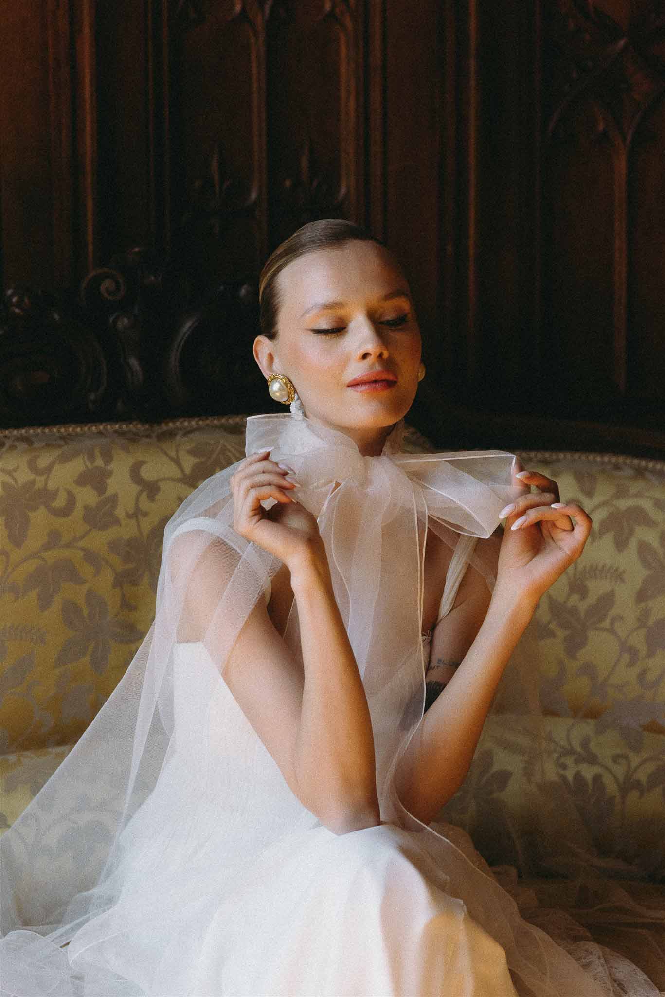 Bride on damask sofa holding sheer organza cape collar with gold pearl drop earrings in panelled room