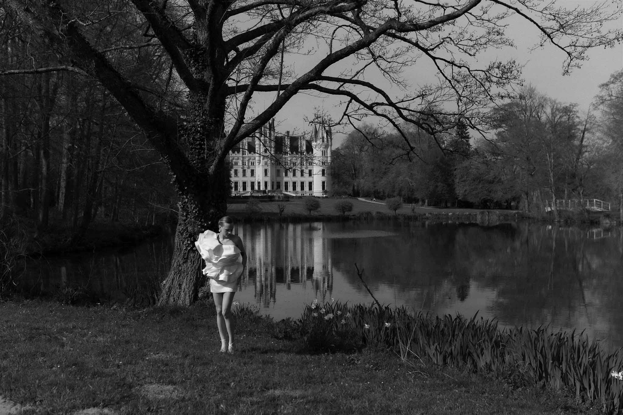 Bride in sculptural ruffled mini dress standing by lake with chateau reflected on water in B&W