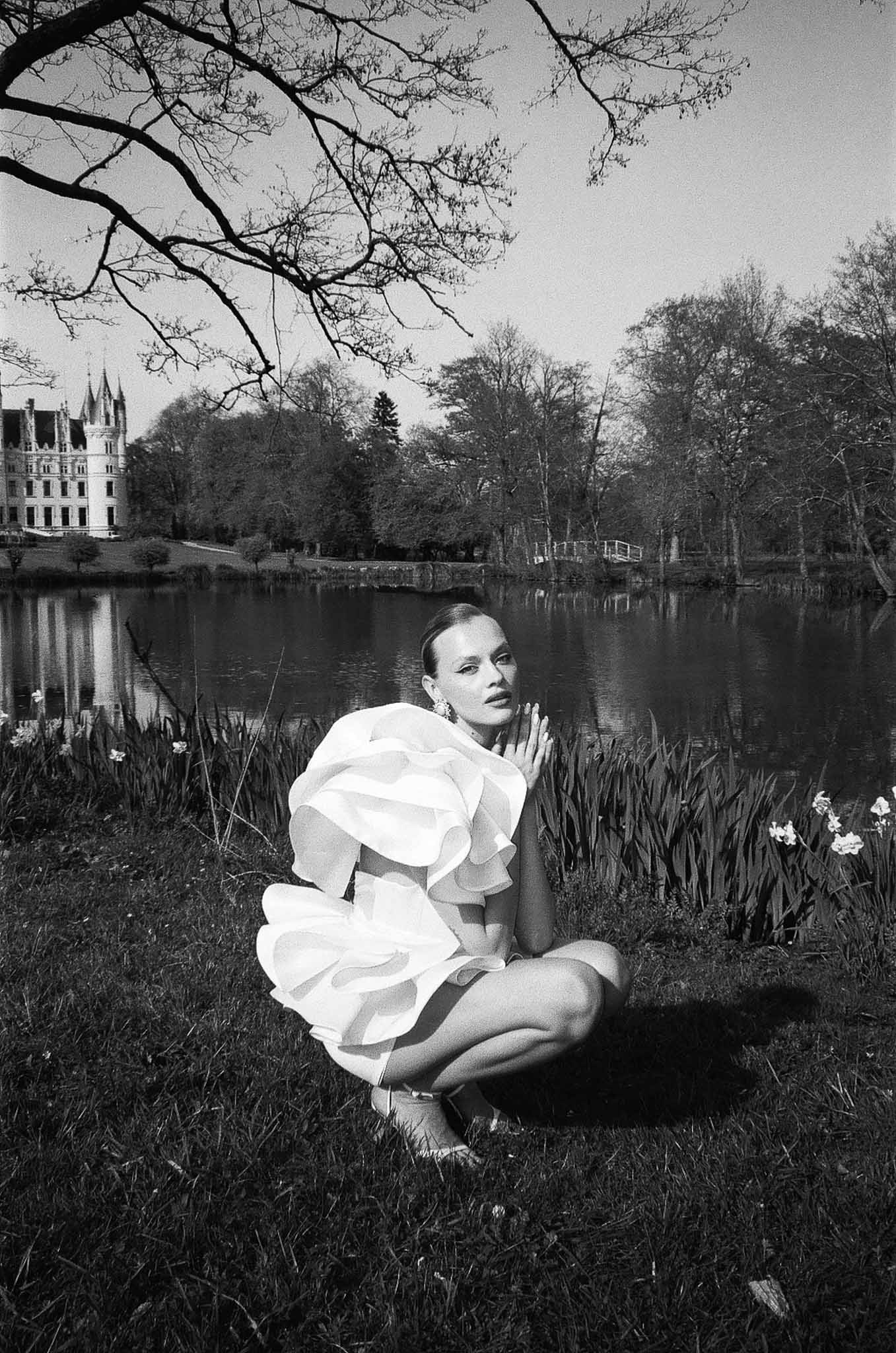 Black and white portrait of bride in ruffled mini dress seated by ornamental lake with turreted chateau in background