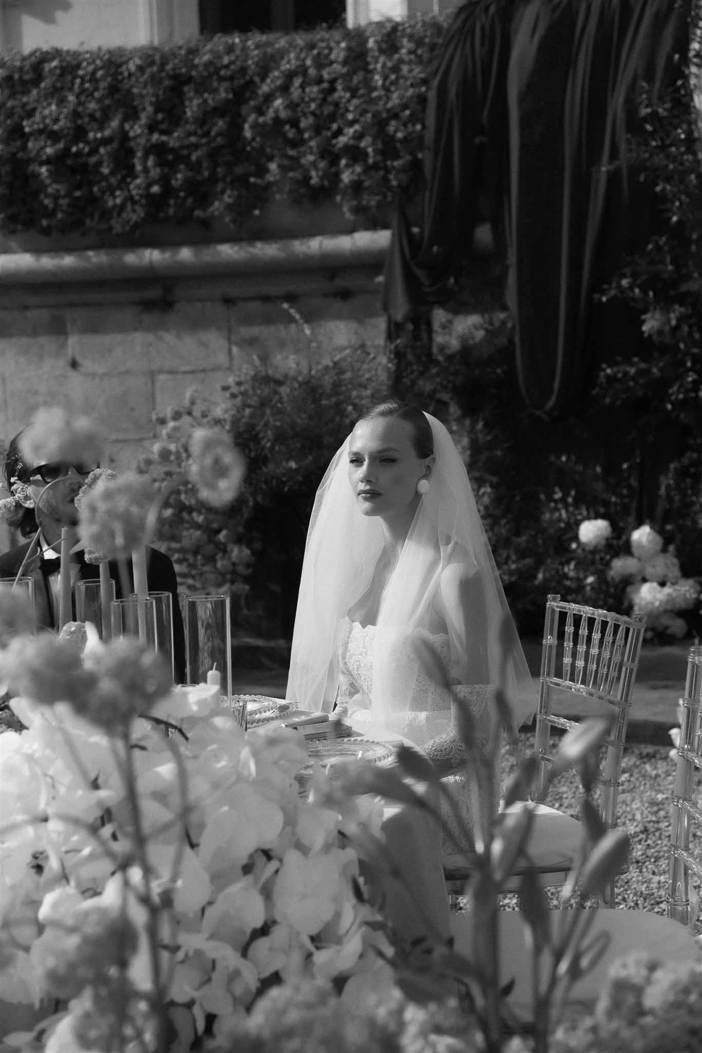 Bride in lace dress and cathedral veil seated at orchid-filled reception table in chateau courtyard in B&W