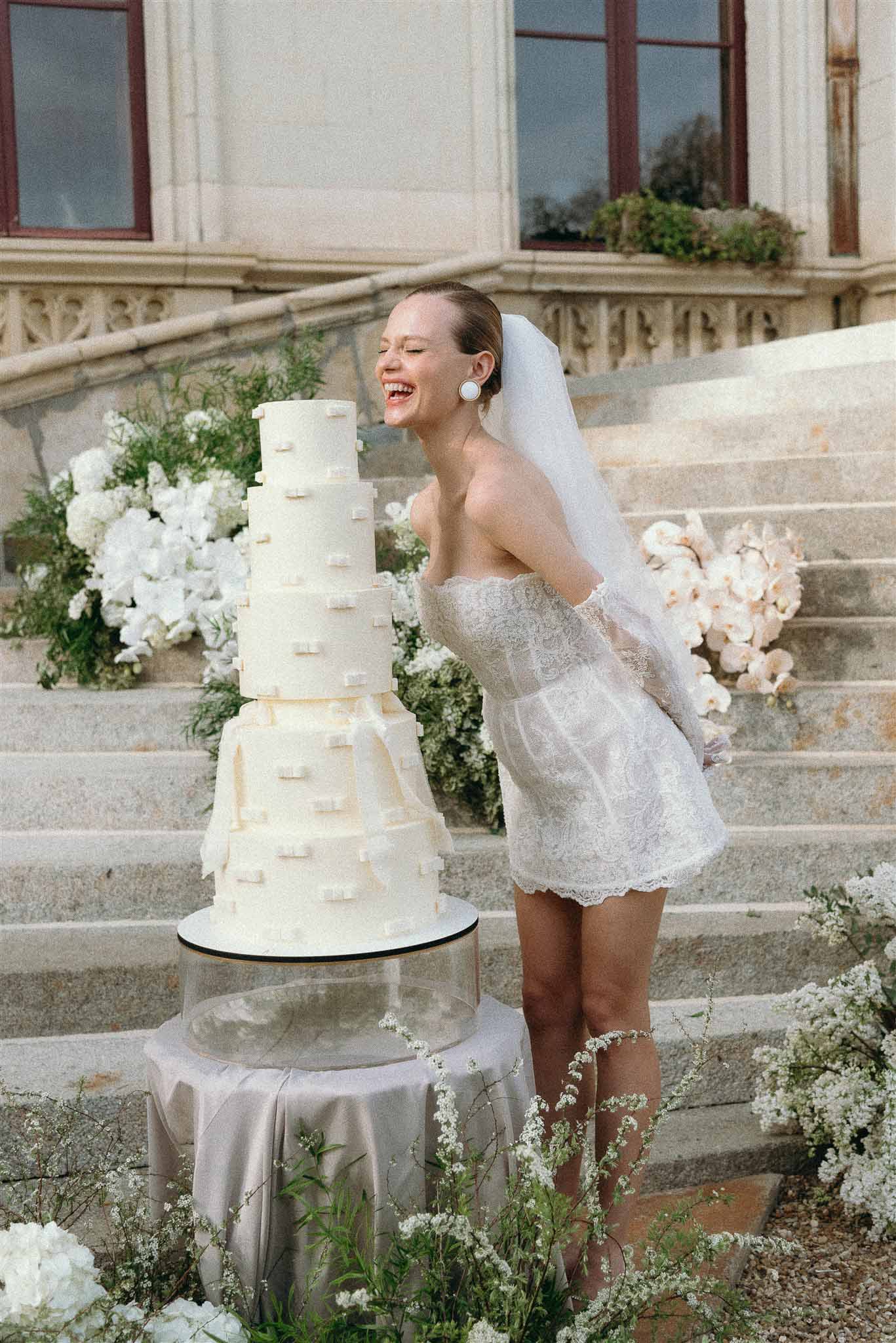Bride in short lace mini dress laughing beside five-tier ivory cake with hydrangea arrangements