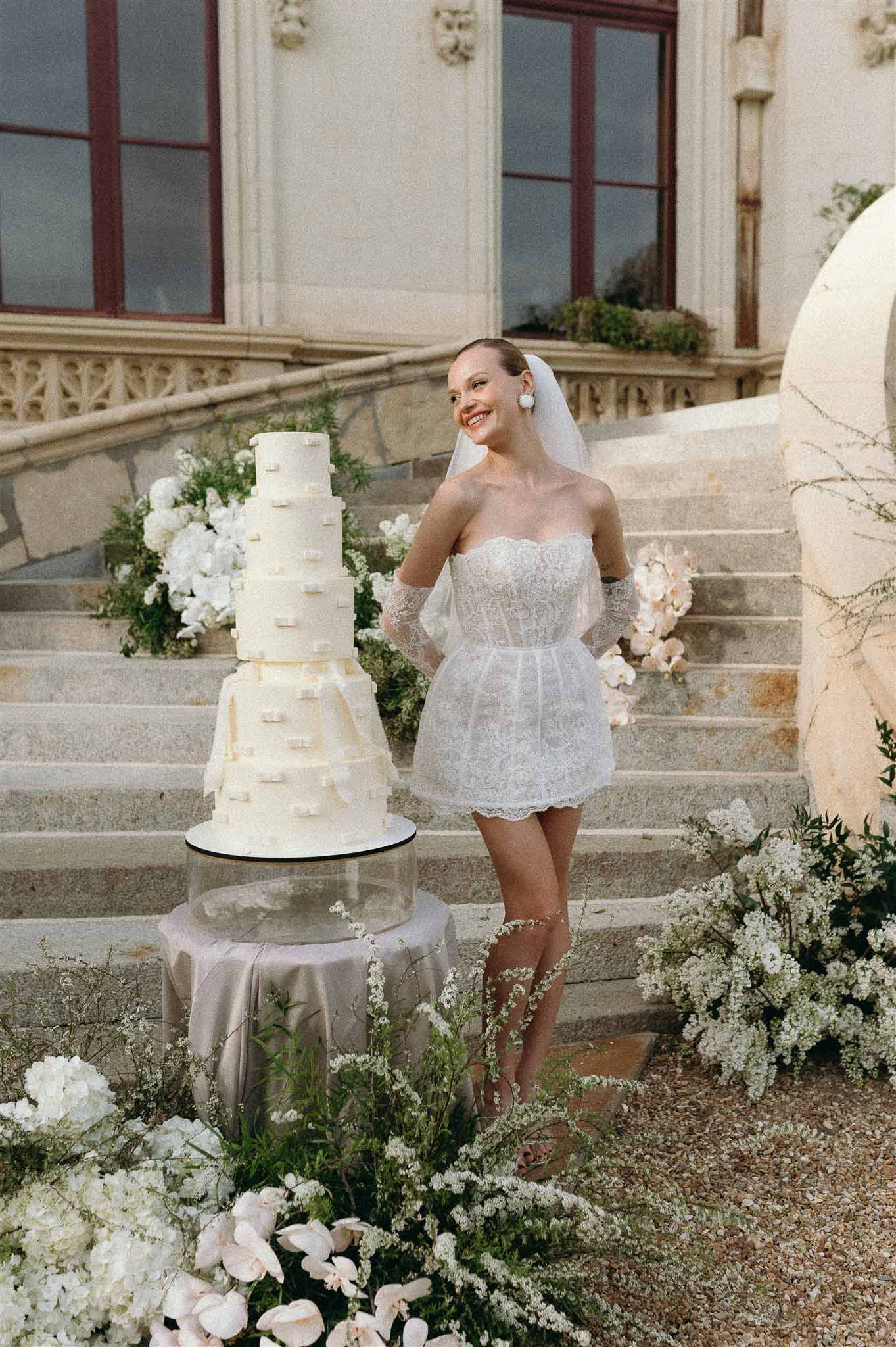 Bride in white lace mini dress and cathedral veil beside five-tier wedding cake on chateau stone steps with white florals