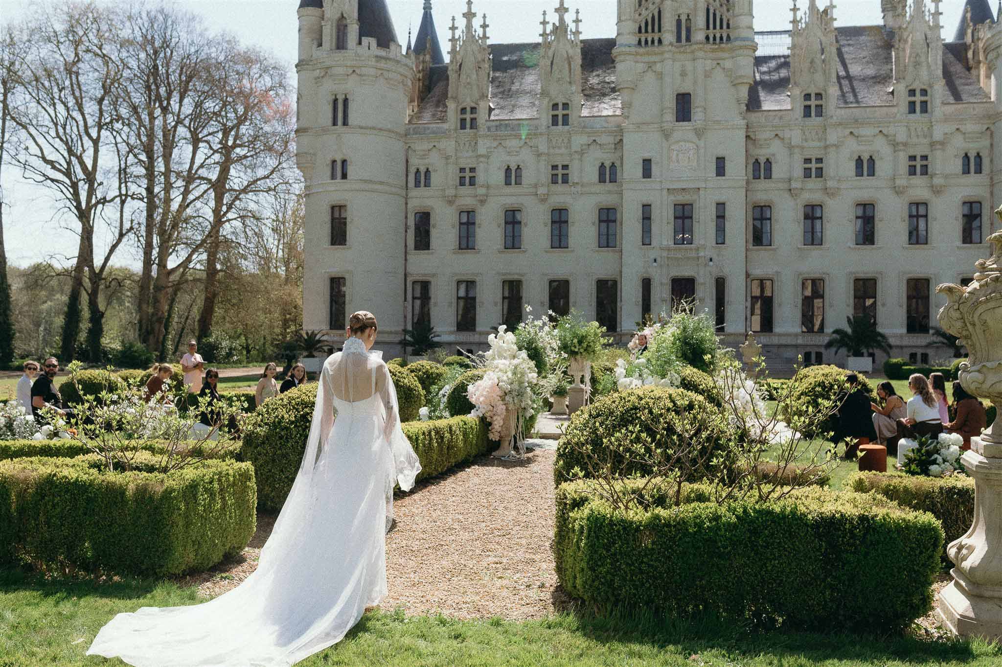 Bride walking down gravel aisle toward flower-decorated altar in formal garden of Gothic Revival chateau
