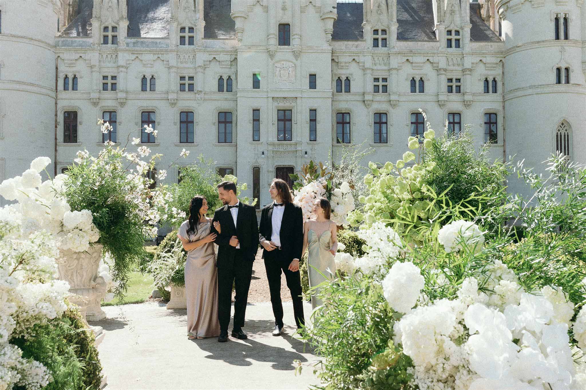 Two couples pose on path flanked by white hydrangea urns before Renaissance chateau limestone facade