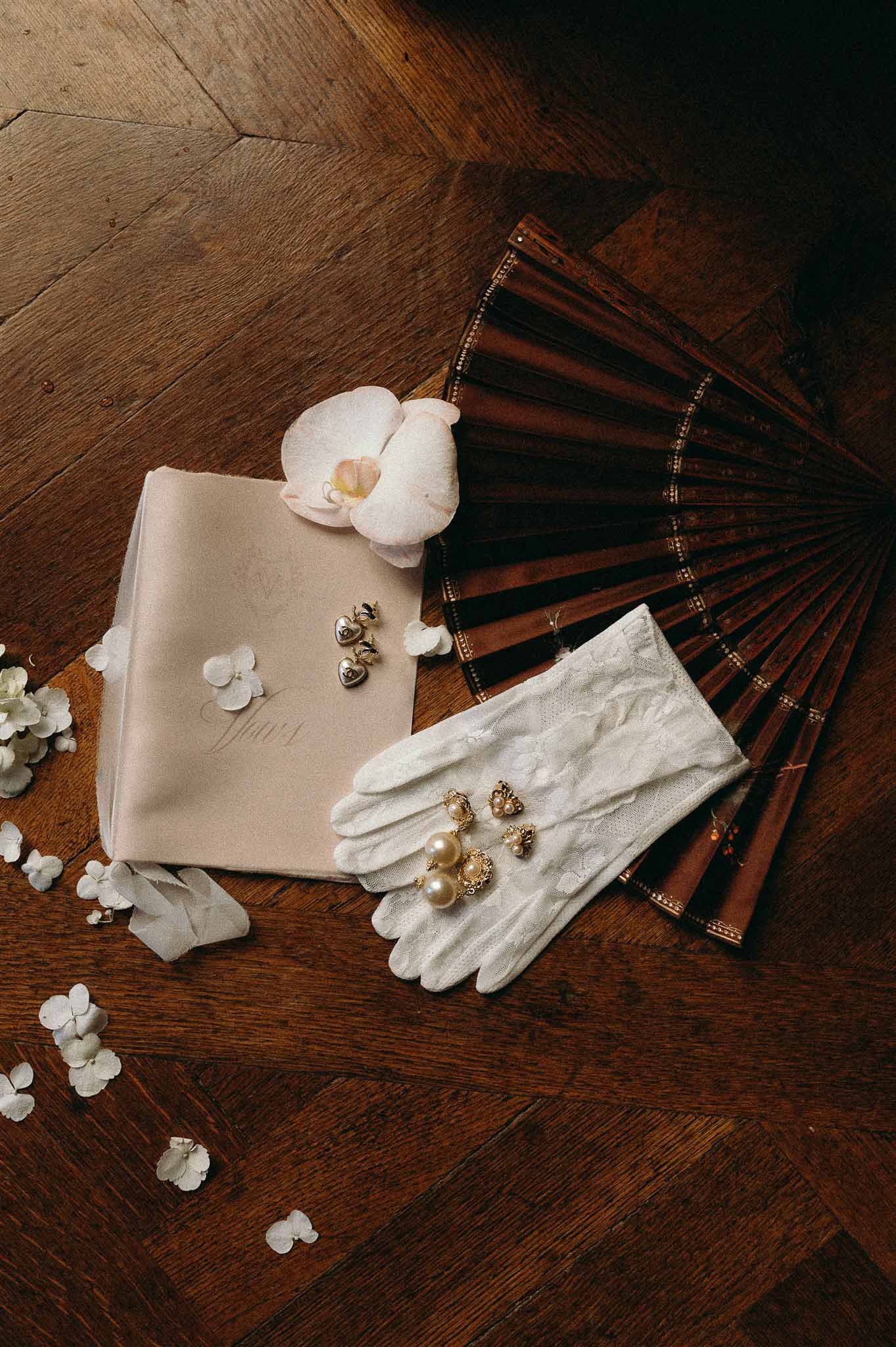 Bridal flat lay with cream vow booklet, lace gloves, mahogany hand fan, gold pearl earrings, and white orchid