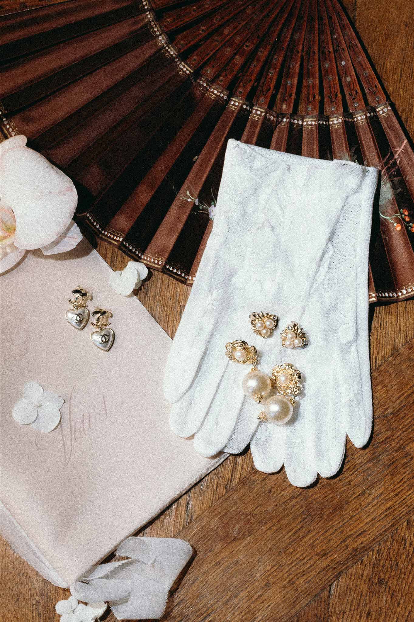 Bridal flat lay on wooden floor with lace gloves, pearl drop earrings, gold heart earrings, and blush stationery card
