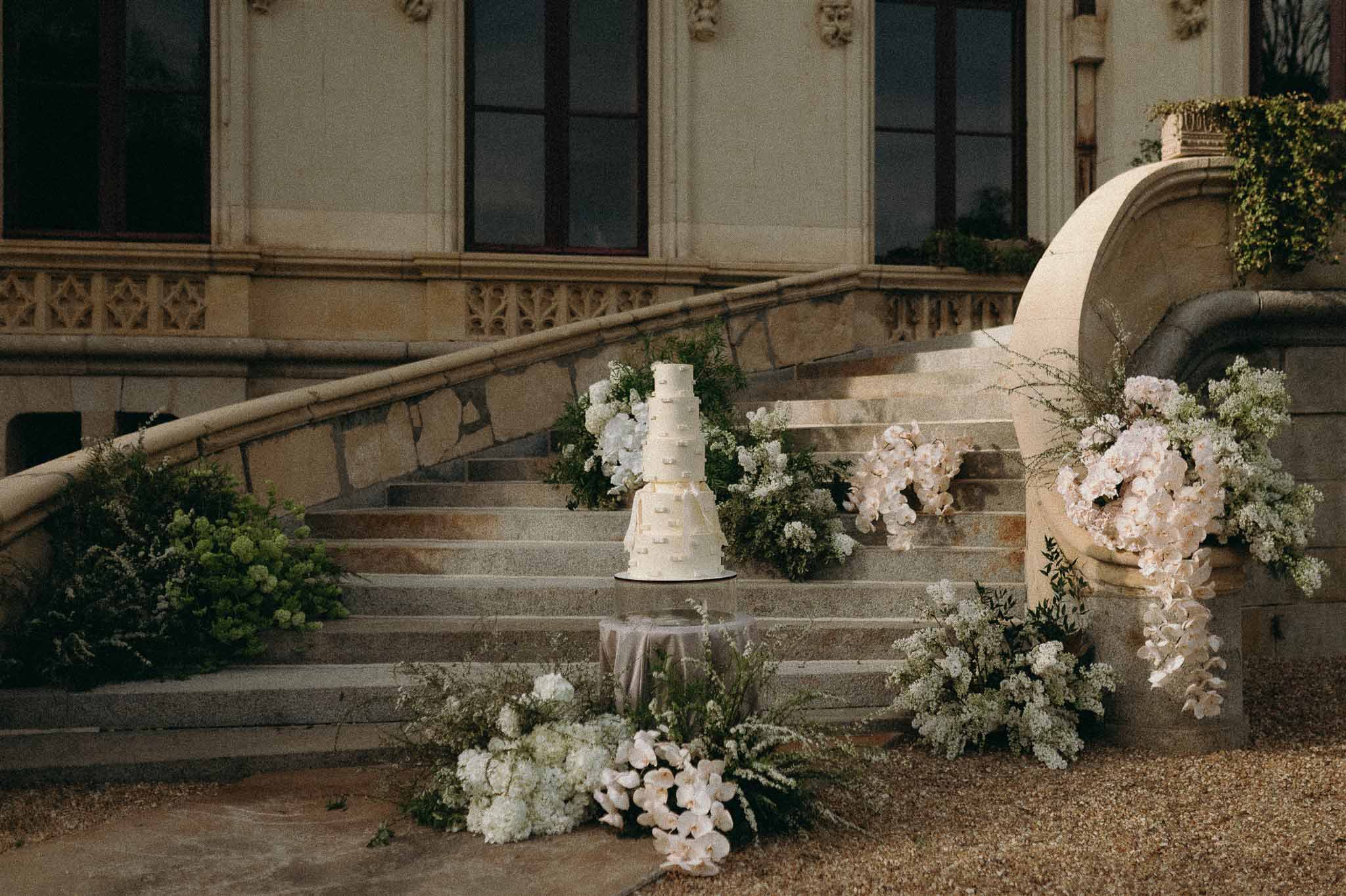 Five-tier ivory buttercream cake surrounded by white orchid and hydrangea arrangements on chateau stone staircase