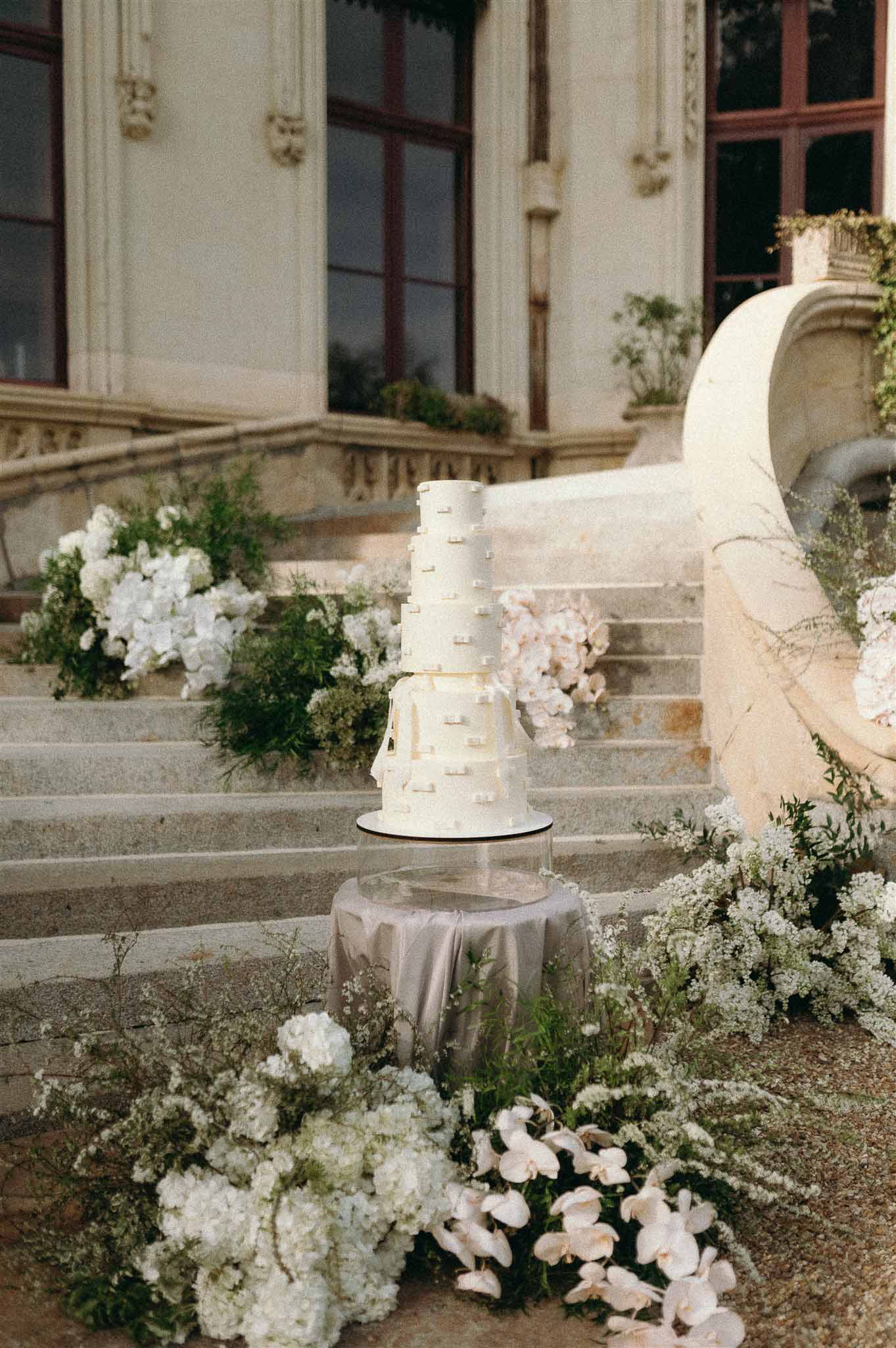 Four-tier ivory cake on acrylic pedestal surrounded by white orchid and hydrangea installations on chateau staircase