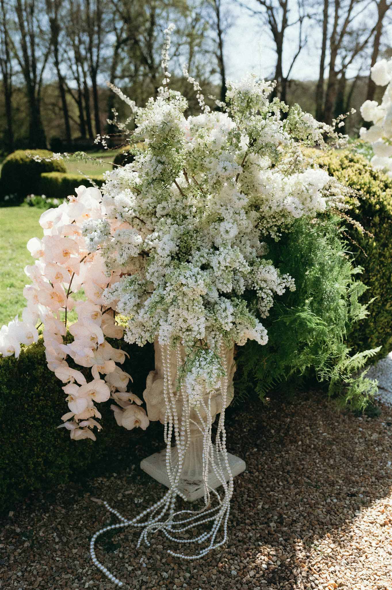 White lilac and cherry blossom urn arrangement with cascading pearl garlands on stone pedestal in formal garden