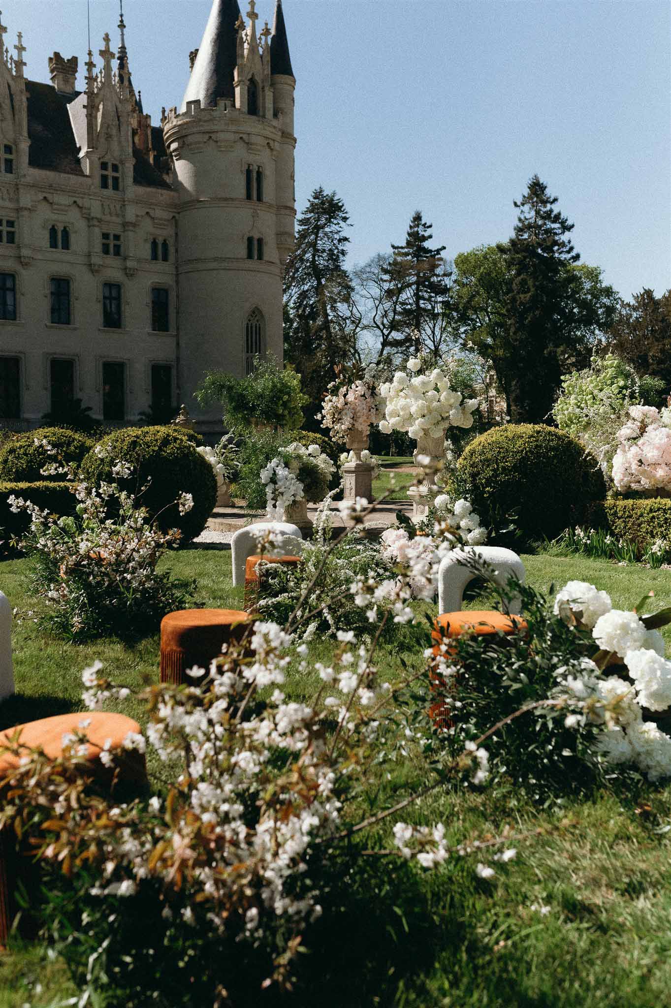 Gothic chateau garden with white hydrangea urns, bouclé chairs, and orange velvet ottomans under cherry blossoms