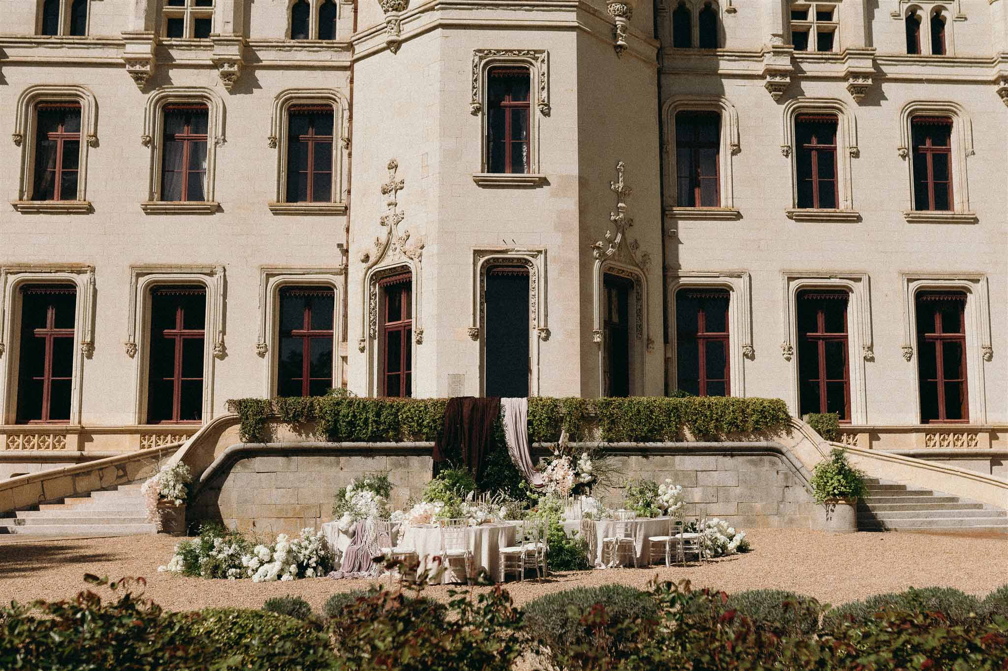 Long reception table with white linen and clear chairs set on chateau courtyard with white hydrangea borders and mauve dra...