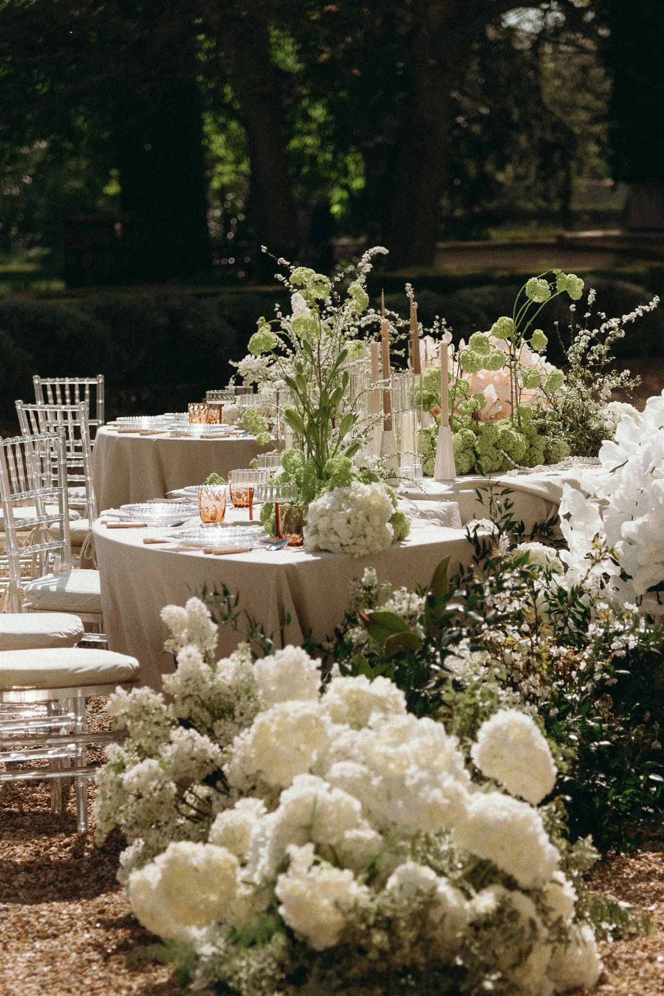 Round reception table with ghost chairs and white hydrangea centerpieces surrounded by ground-level floral displays