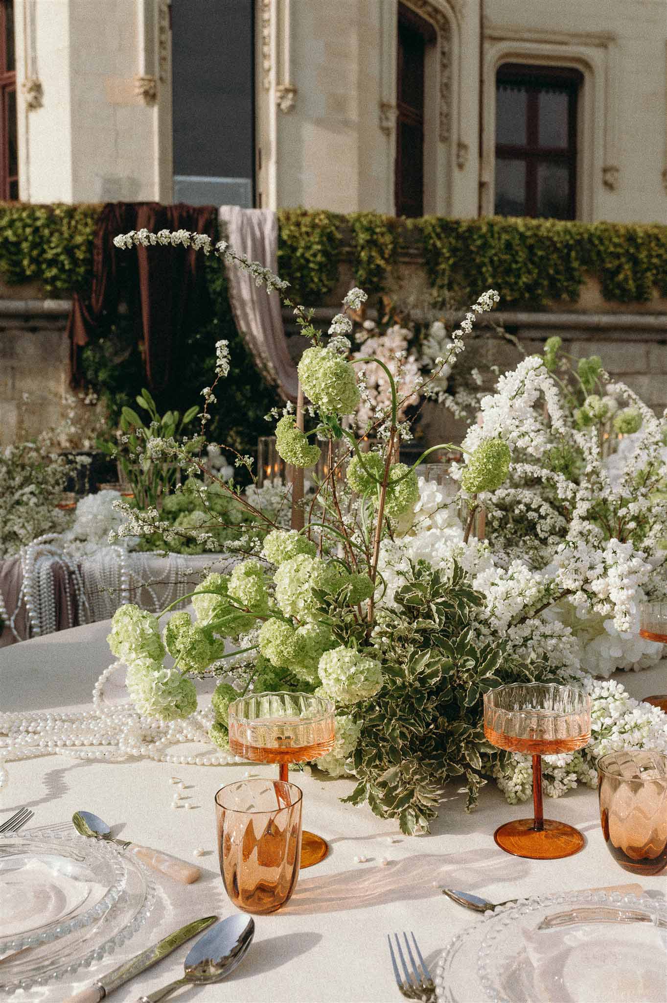 Outdoor reception table with green viburnum centerpiece, amber coupe glasses, and silver cutlery at a French chateau