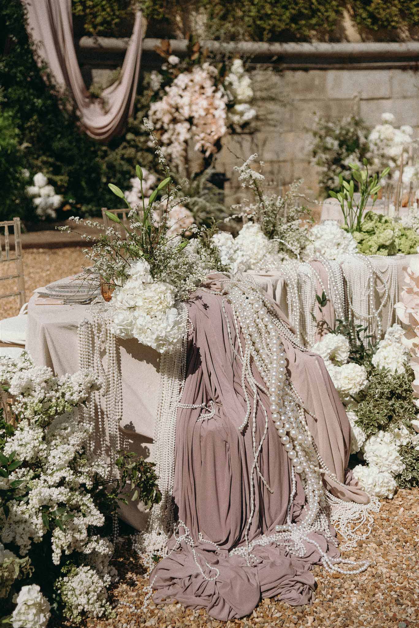 Reception table with dusty mauve linen, pearl garlands, white hydrangea runner and amber glass vases against stone wall