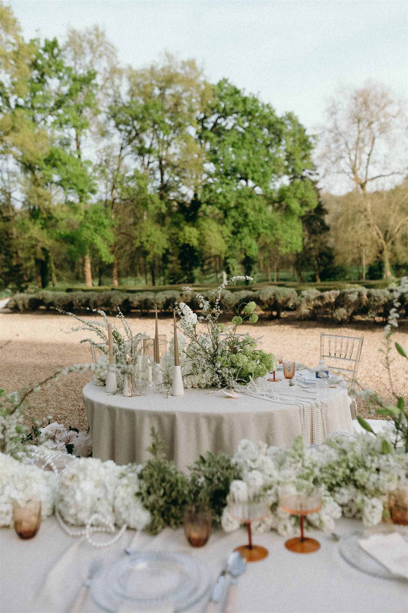 Round table with white hydrangea and viburnum centerpiece, pearl strands, and amber glassware in garden