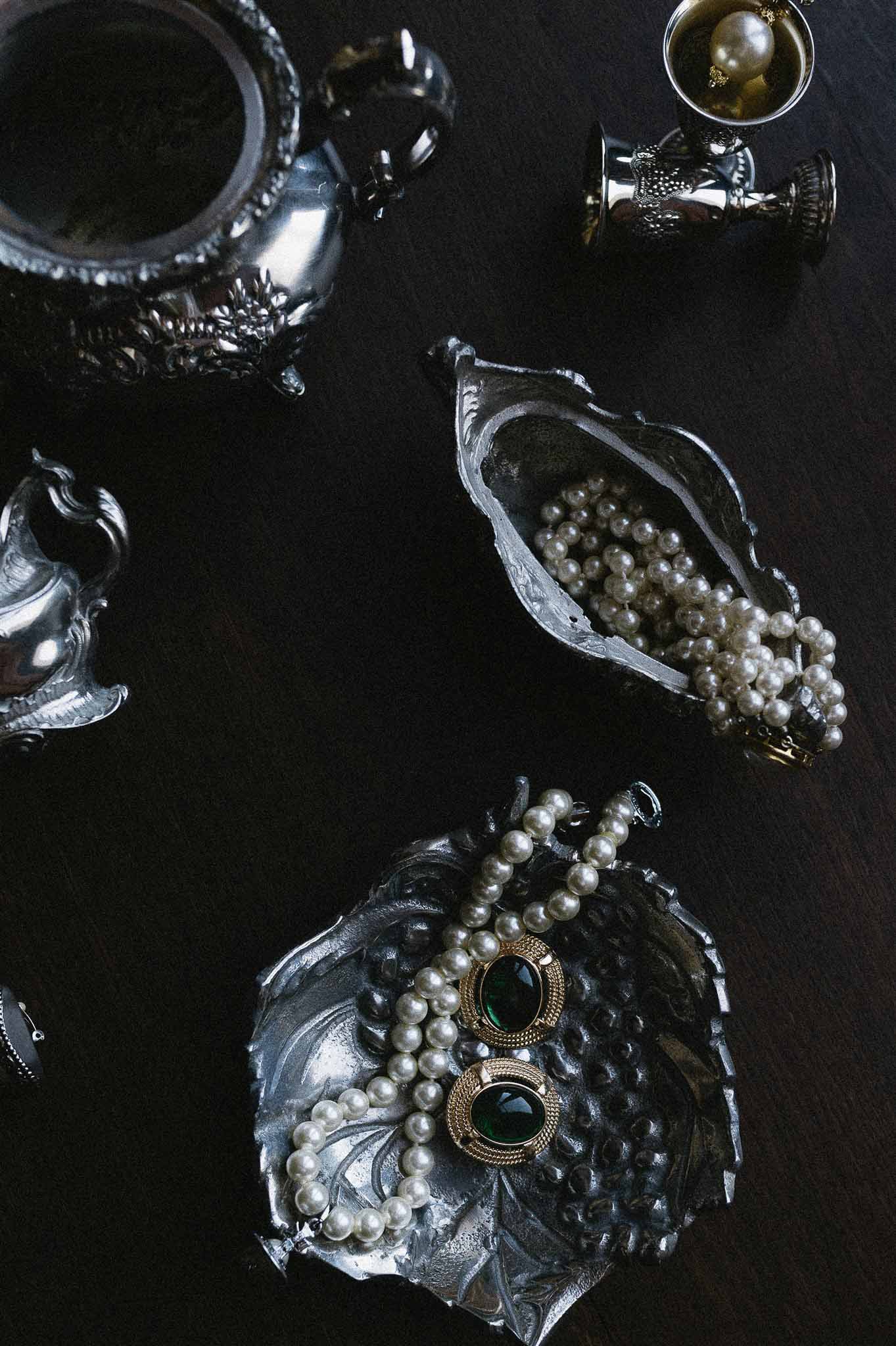 Antique silver vessels with pearl necklaces and emerald green brooches on dark surface