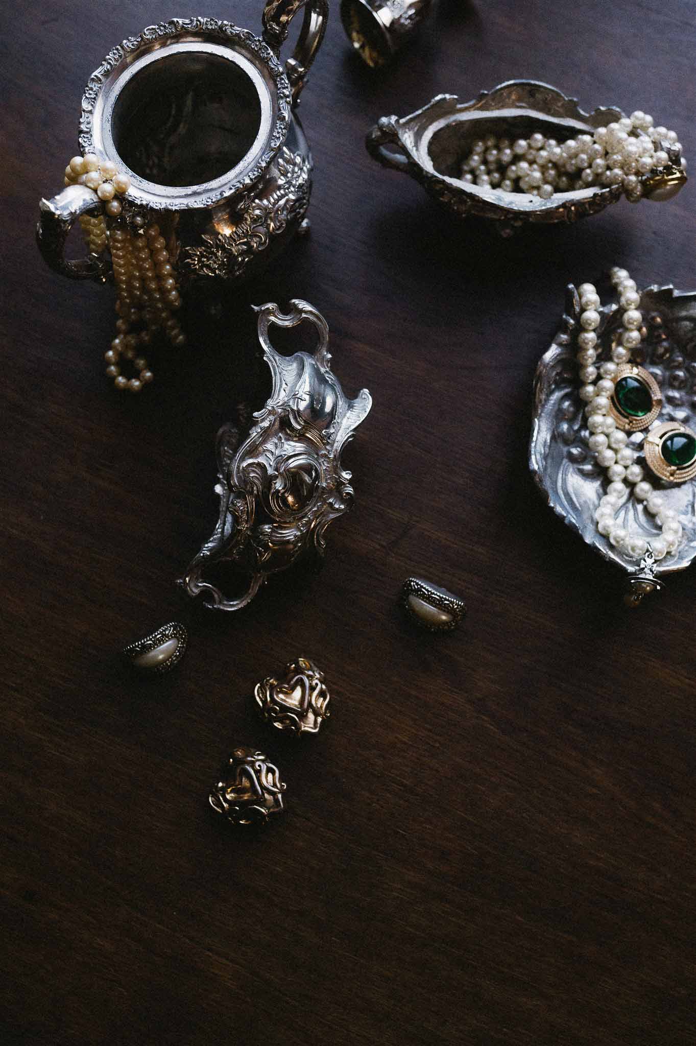 Vintage jewelry flat lay with pearl necklaces gold emerald brooches silver vessels and marcasite rings on dark wood