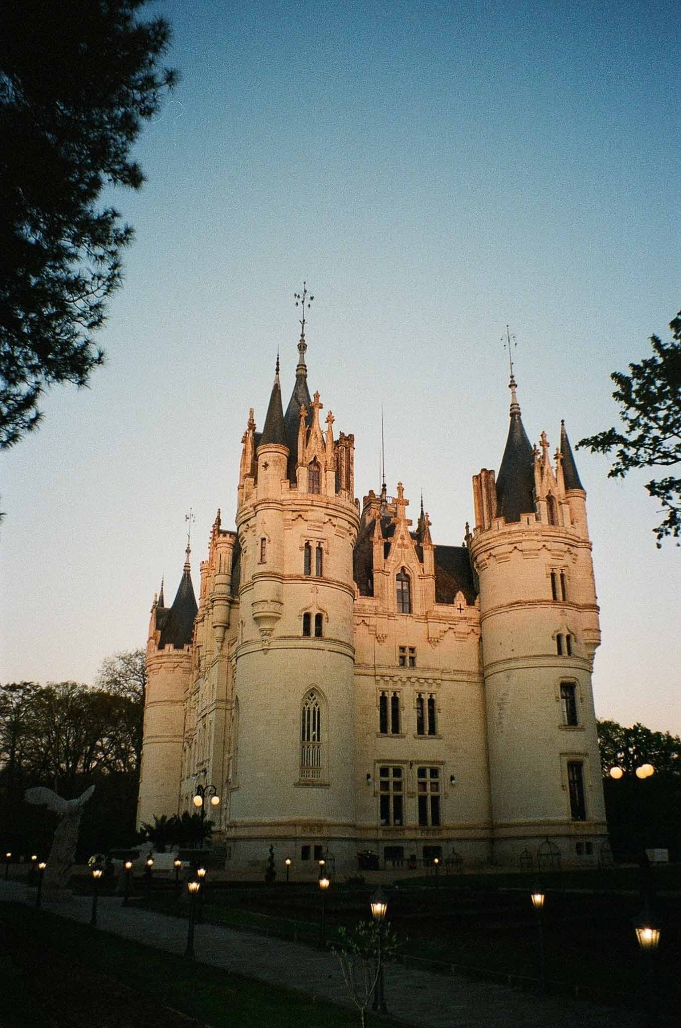 Gothic-Renaissance French chateau with conical turrets and ornate stone detailing lit by lantern post lights at dusk