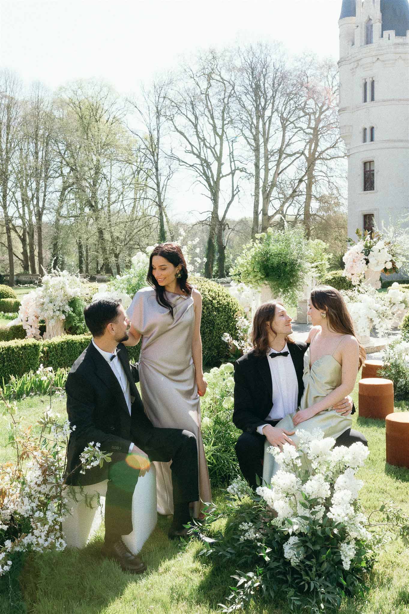 Four guests in tuxedos and satin dresses among white and blush floral installations in chateau garden cocktail hour