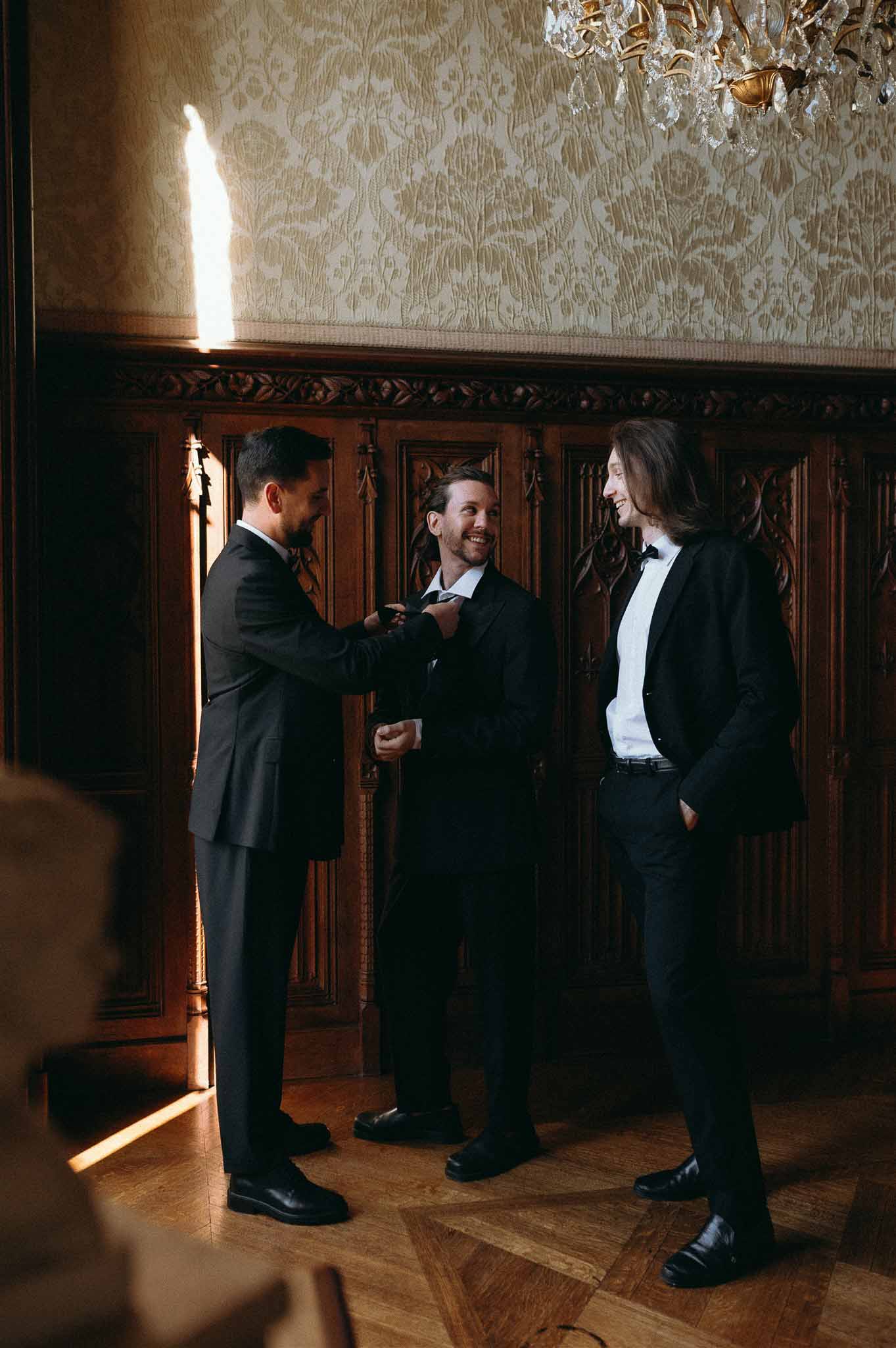 Three groomsmen in black tuxedos laughing while adjusting ties in wood-paneled chateau room