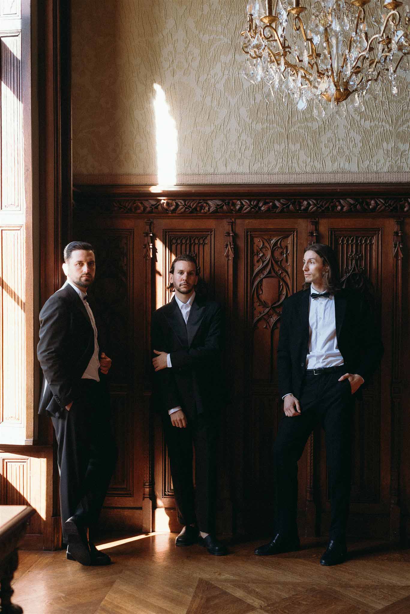 Three groomsmen in black tuxedos posed against carved wood paneling beneath crystal chandelier in chateau interior