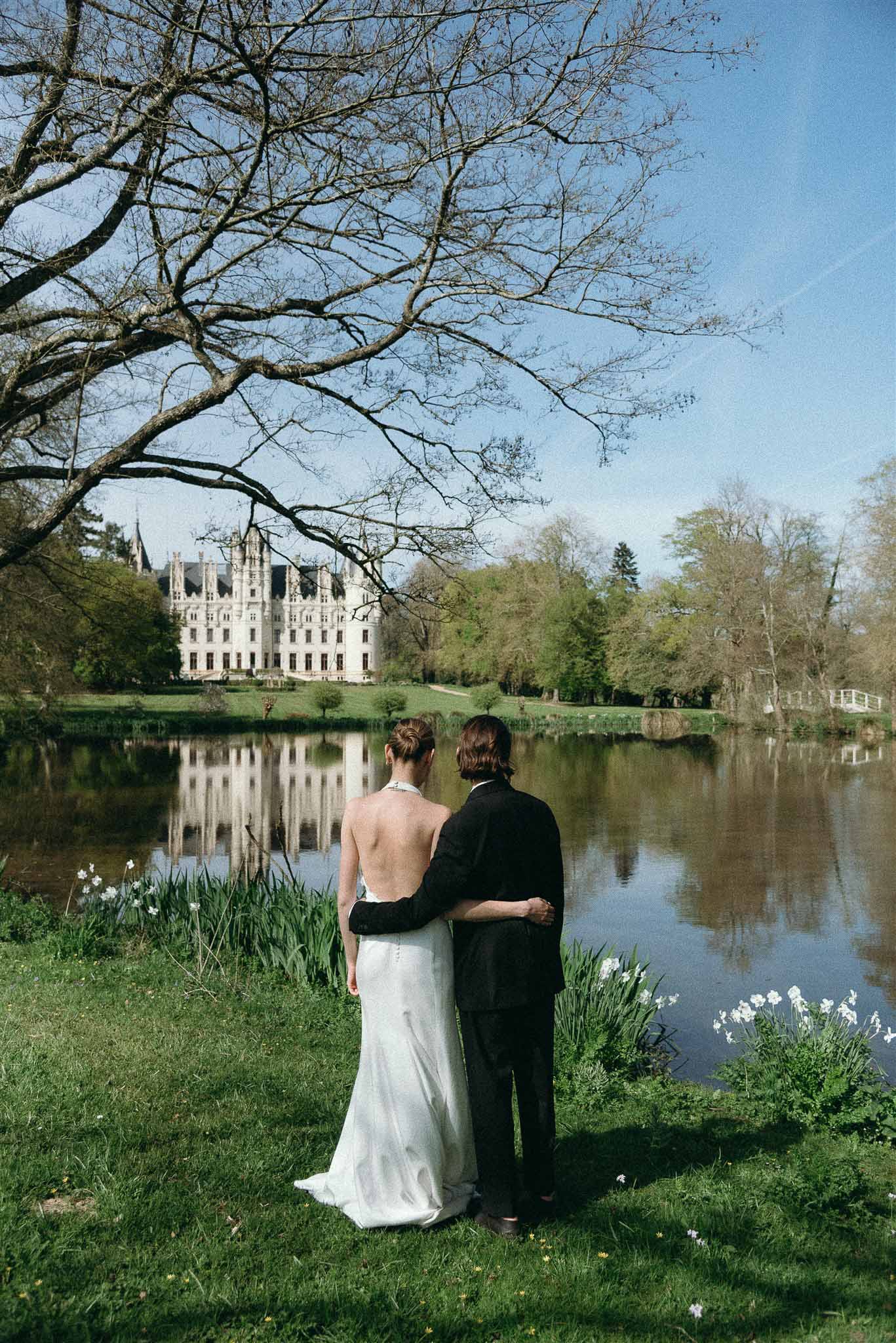 Bride and groom viewed from behind at lakeside with Renaissance chateau reflected in still water