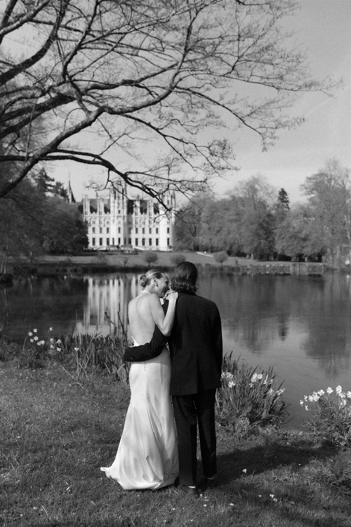 Black and white of couple from behind at lake edge with Gothic chateau reflected in still water