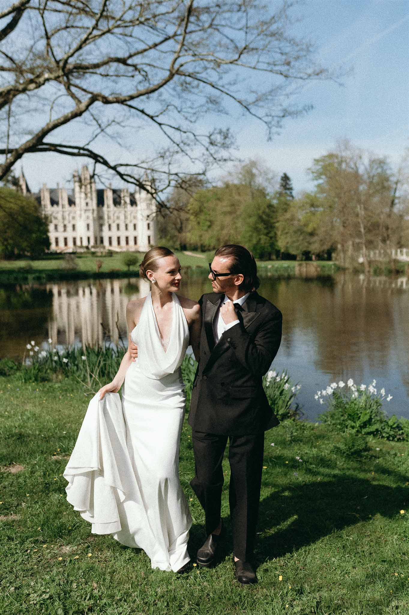Couple on lakeside lawn bride in ivory halter tiered gown groom in black double-breasted tuxedo chateau reflected in water