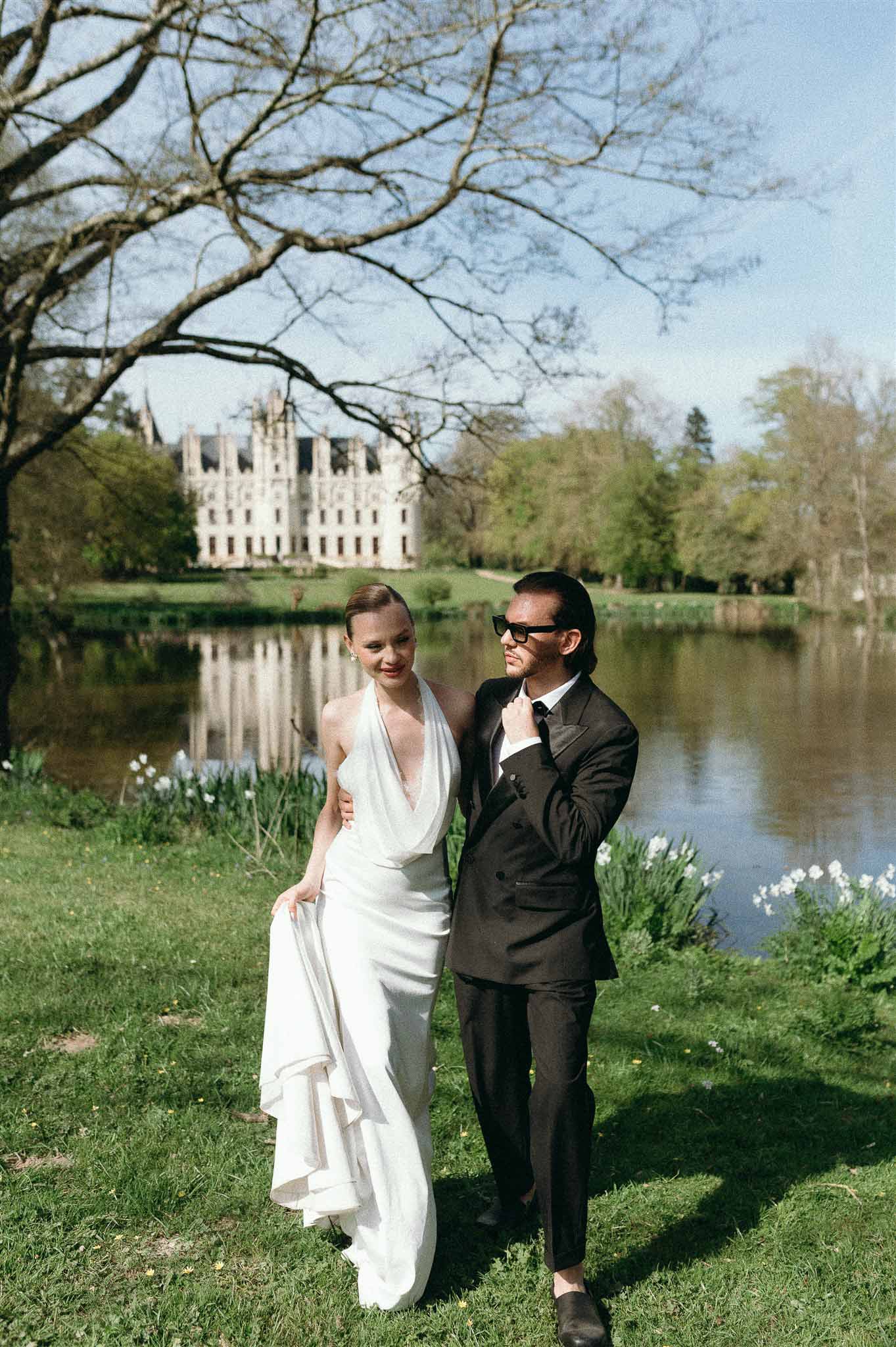 Bride in white halter gown and groom in black suit posing by ornamental lake reflecting French chateau