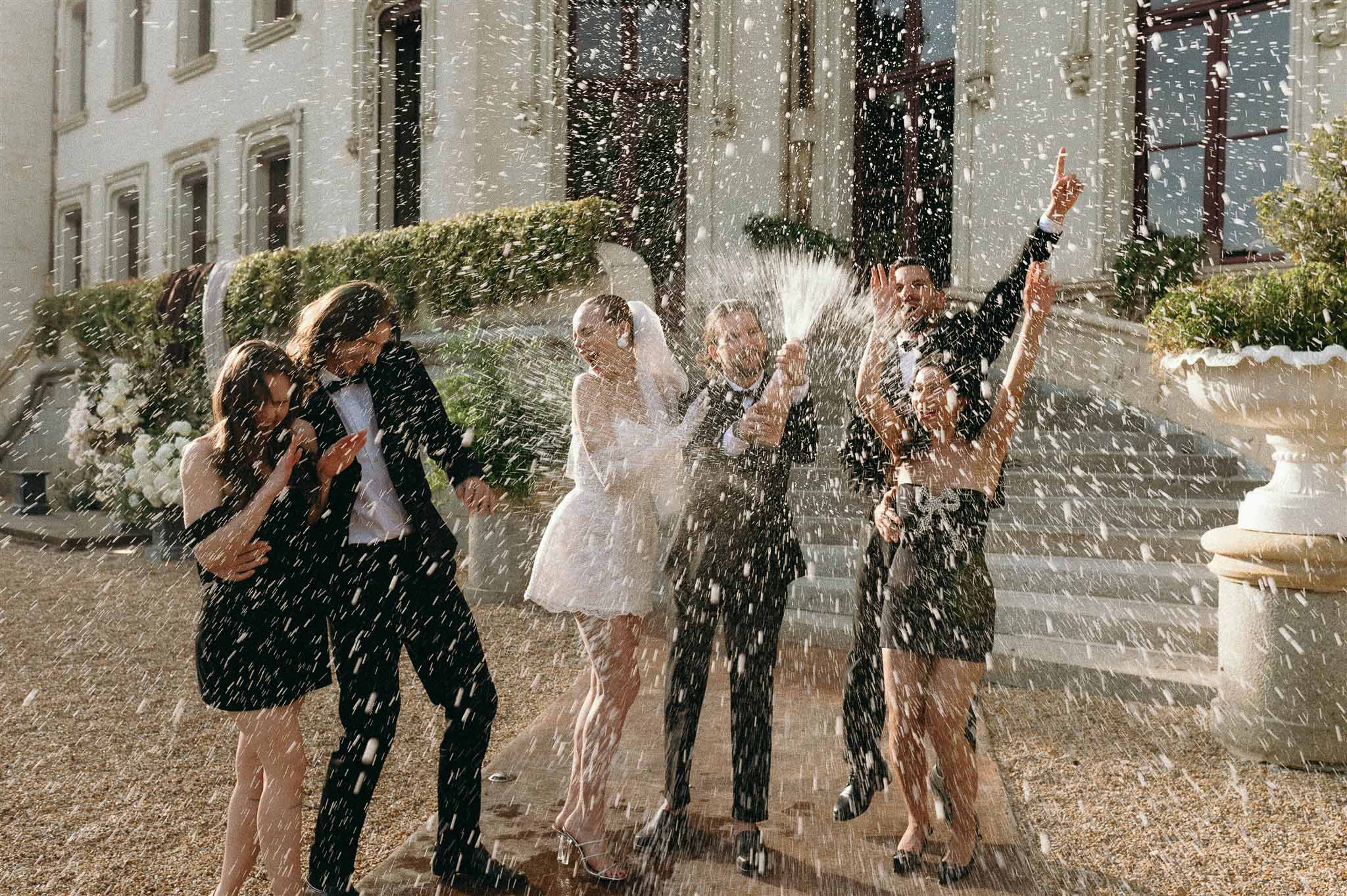 Bride and groom spraying champagne with guests on chateau gravel courtyard in celebration