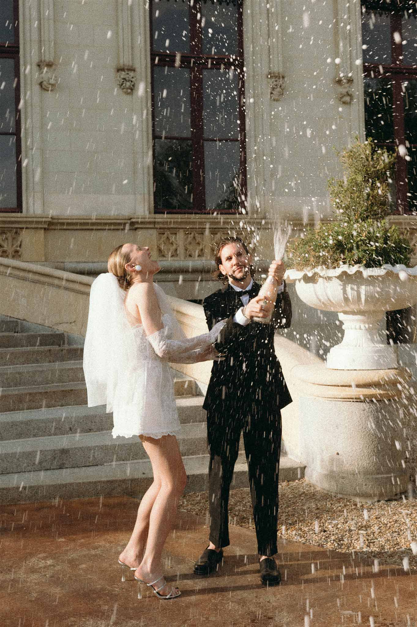 Couple popping champagne on chateau terrace bride in lace mini dress with veil groom in dark suit