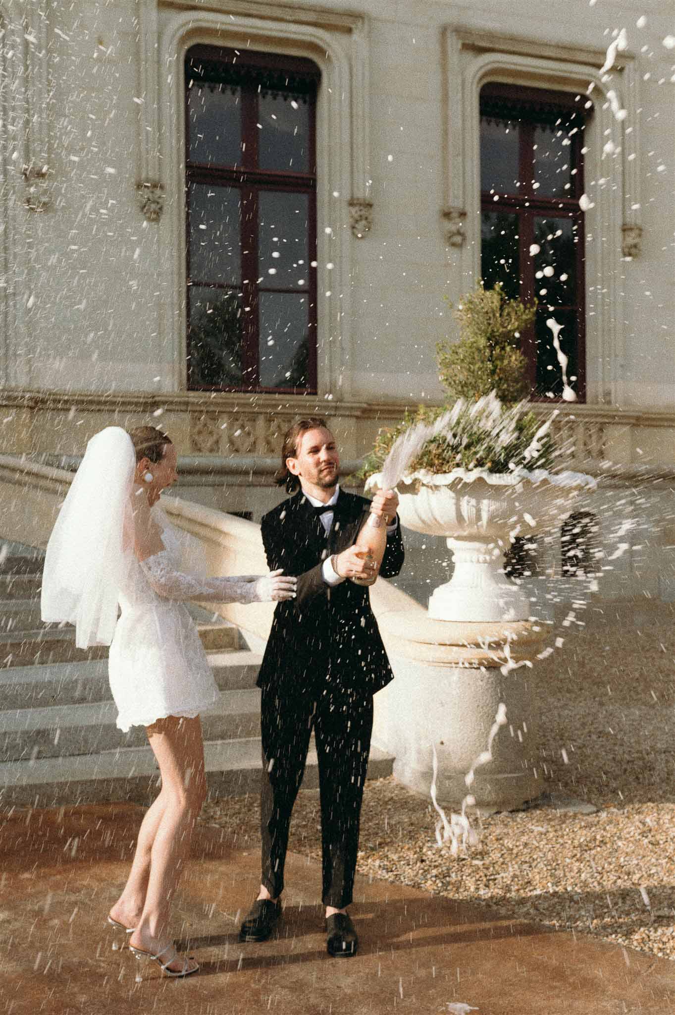 Couple sabering rose champagne on chateau terrace bride in white mini dress with veil groom in black suit