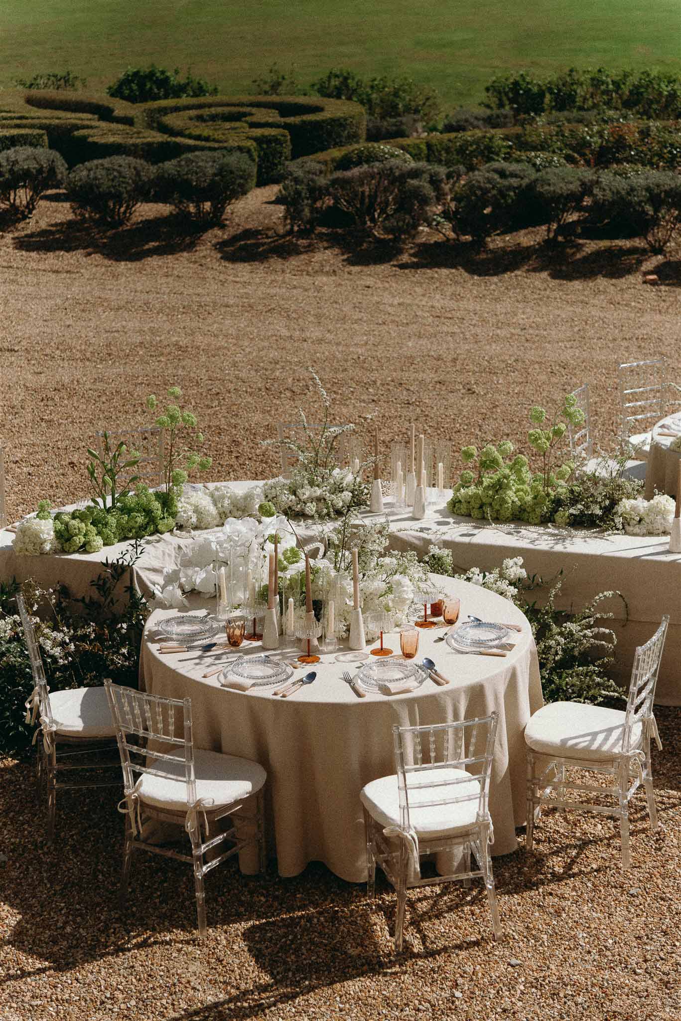 Round reception table with white peonies and hydrangeas centerpiece, acrylic chairs, on gravel terrace with hedge garden