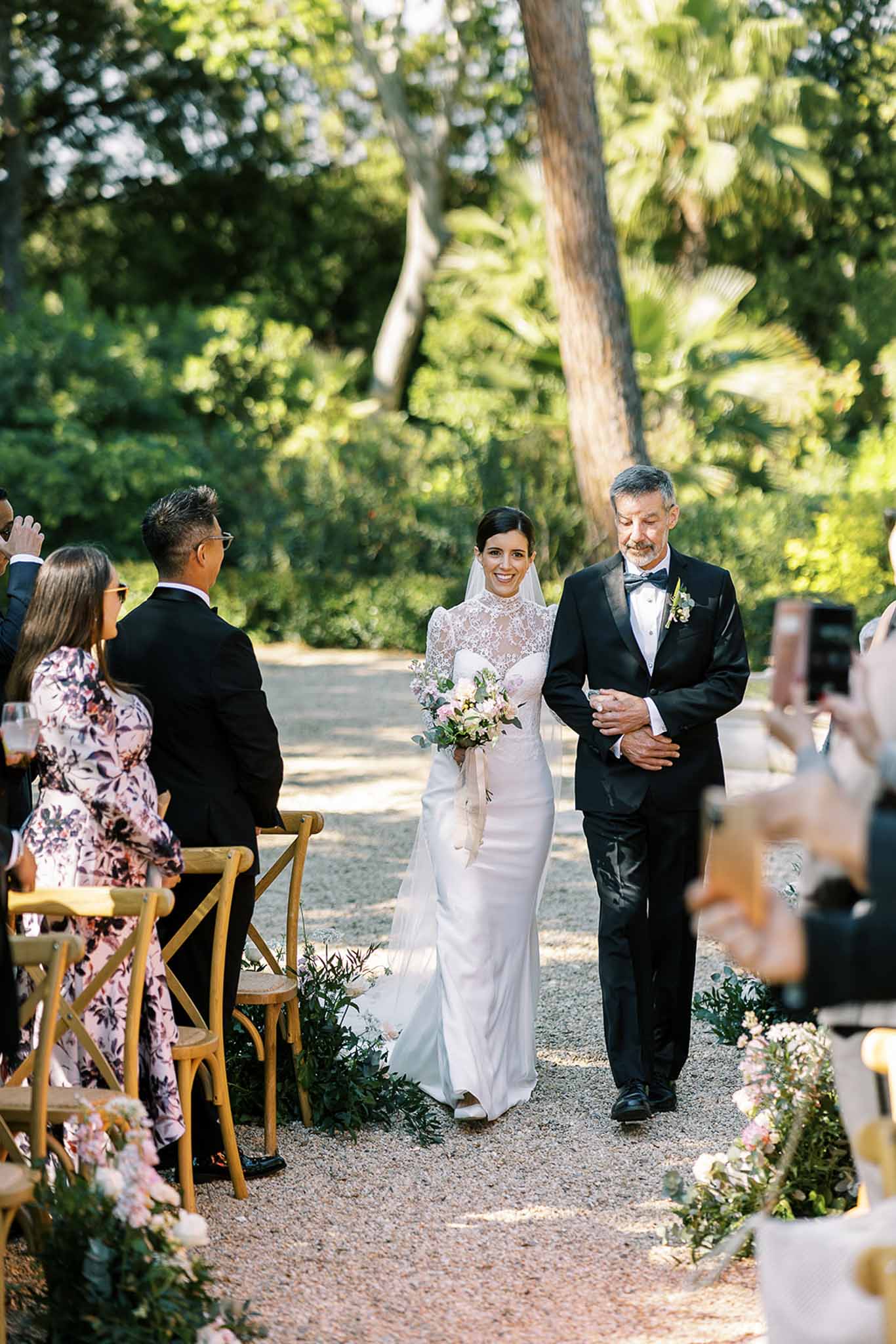Bride in ivory satin gown with lace overlay walks down garden aisle with father past greenery and blush floral arrangements