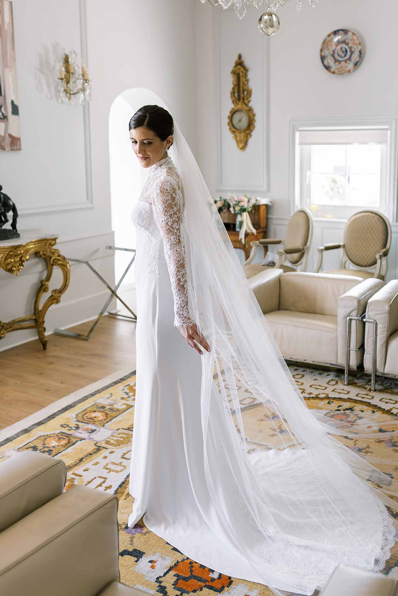 Bride in white lace long-sleeved gown with cathedral veil glancing over shoulder in classical salon with chandelier