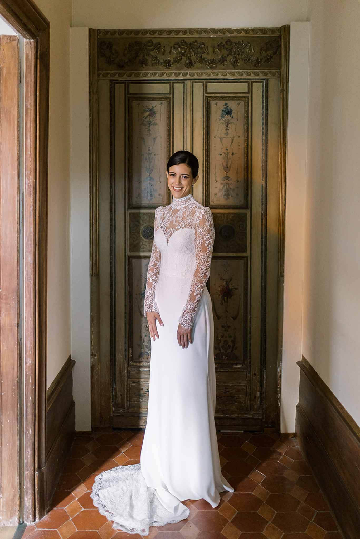 Bride in high-neck lace gown with crepe skirt smiling in front of painted chateau door