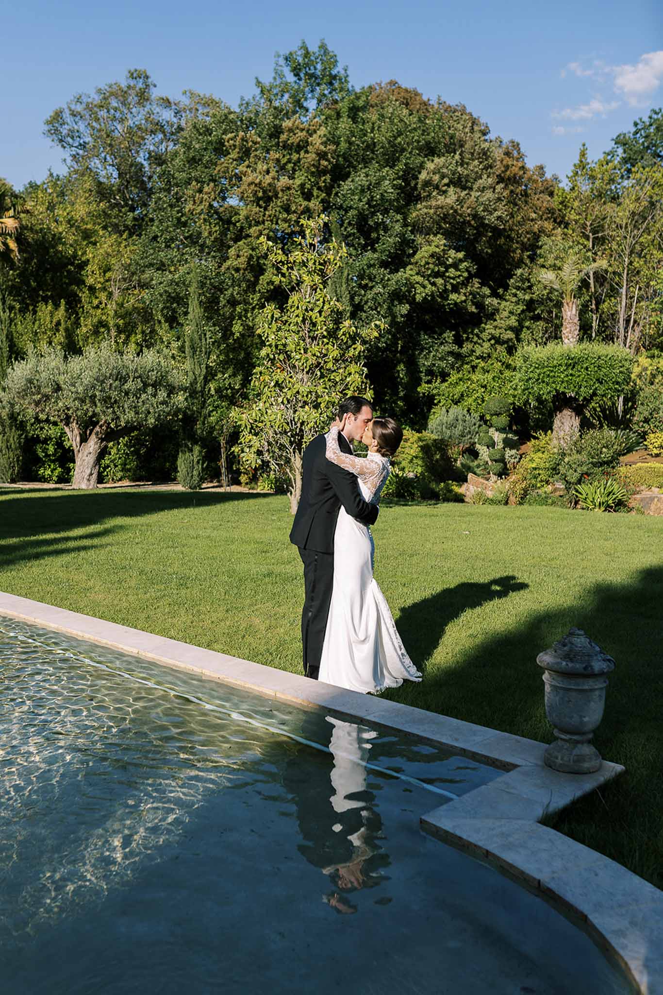 Couple kissing at edge of reflecting pool with stone urn on formal estate lawn with topiary
