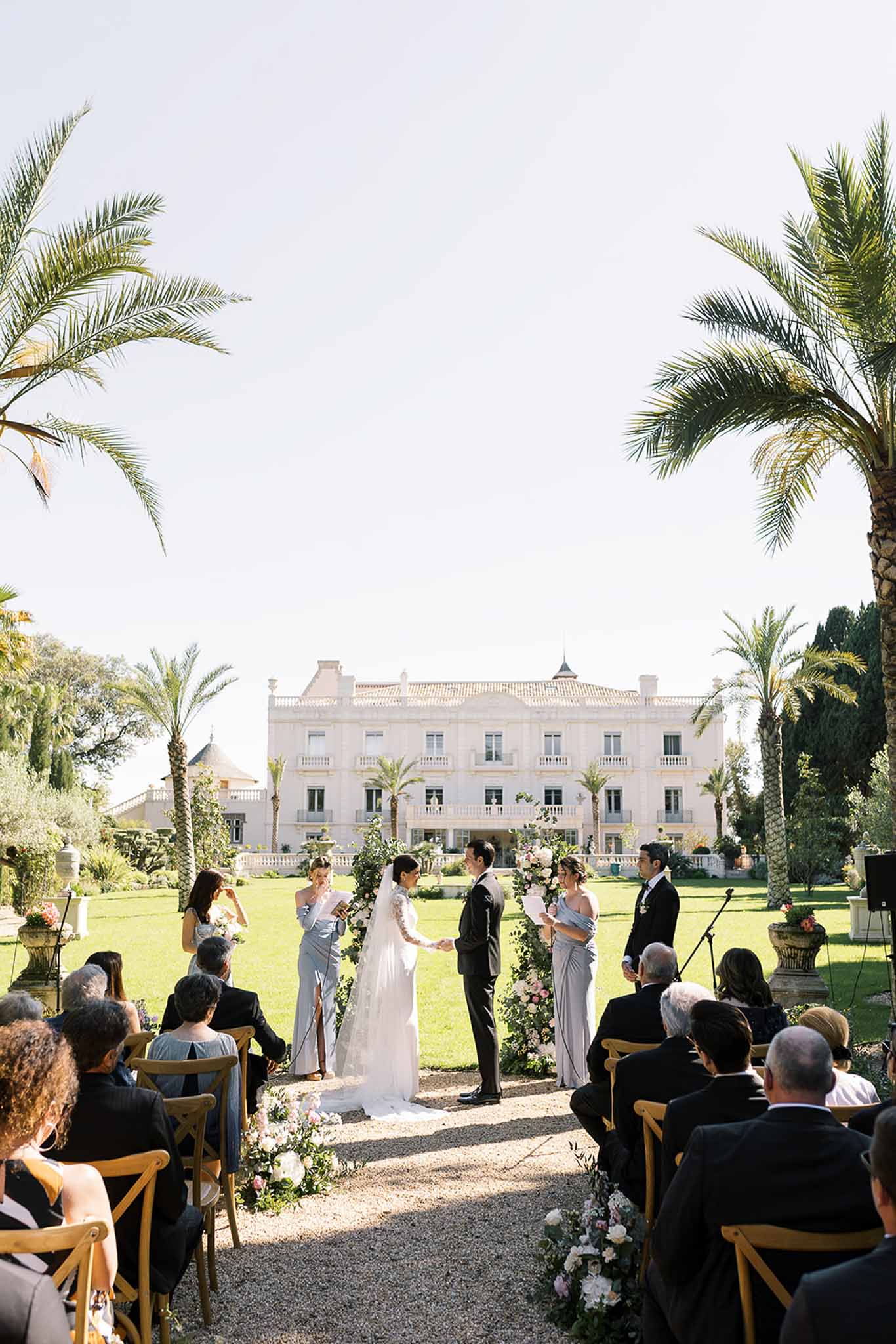 Outdoor wedding ceremony on lawn before Belle Epoque villa with floral arch, seated guests, and bridal party