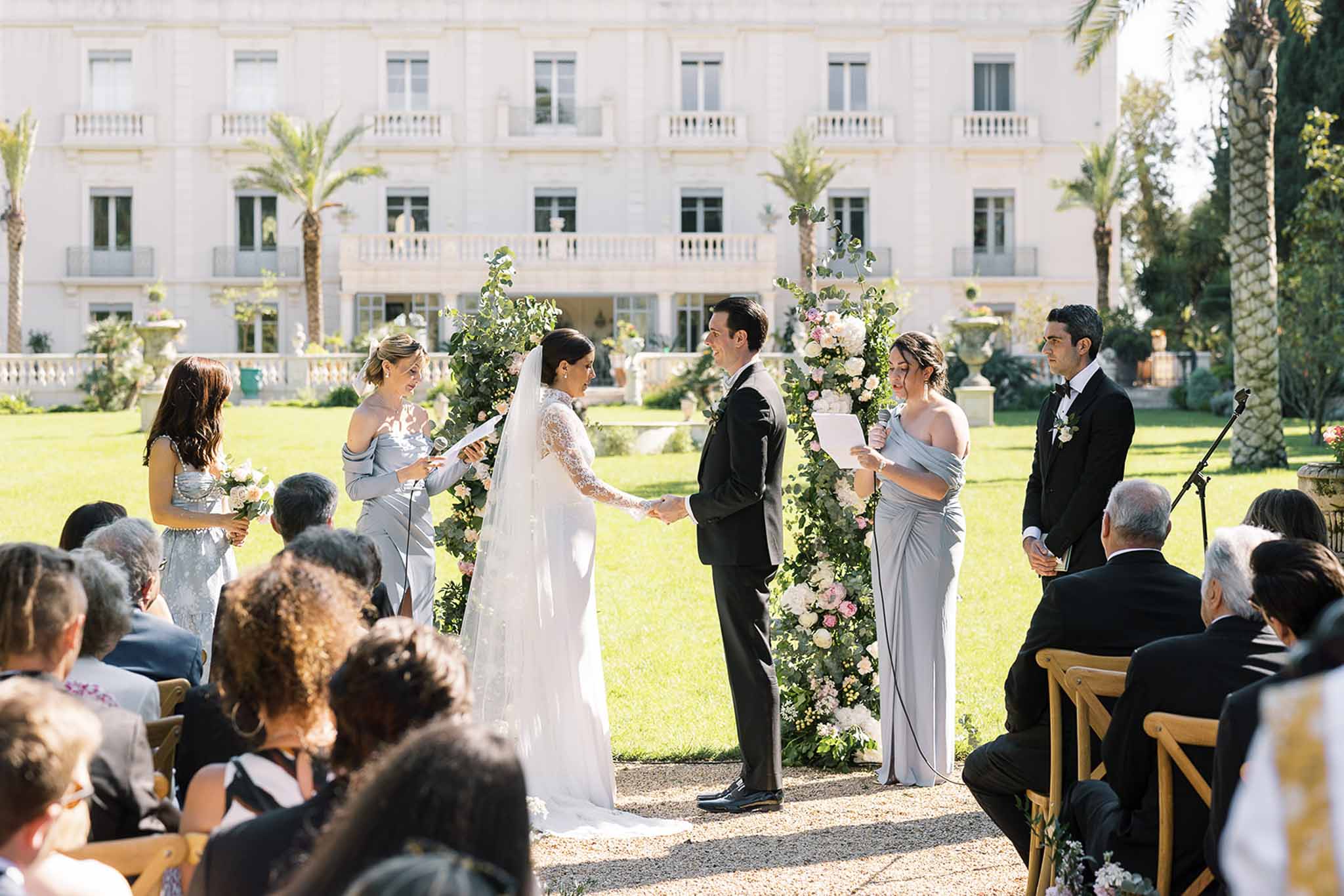 Outdoor ceremony at neoclassical villa with floral arch, grey bridesmaids, and 40 seated guests