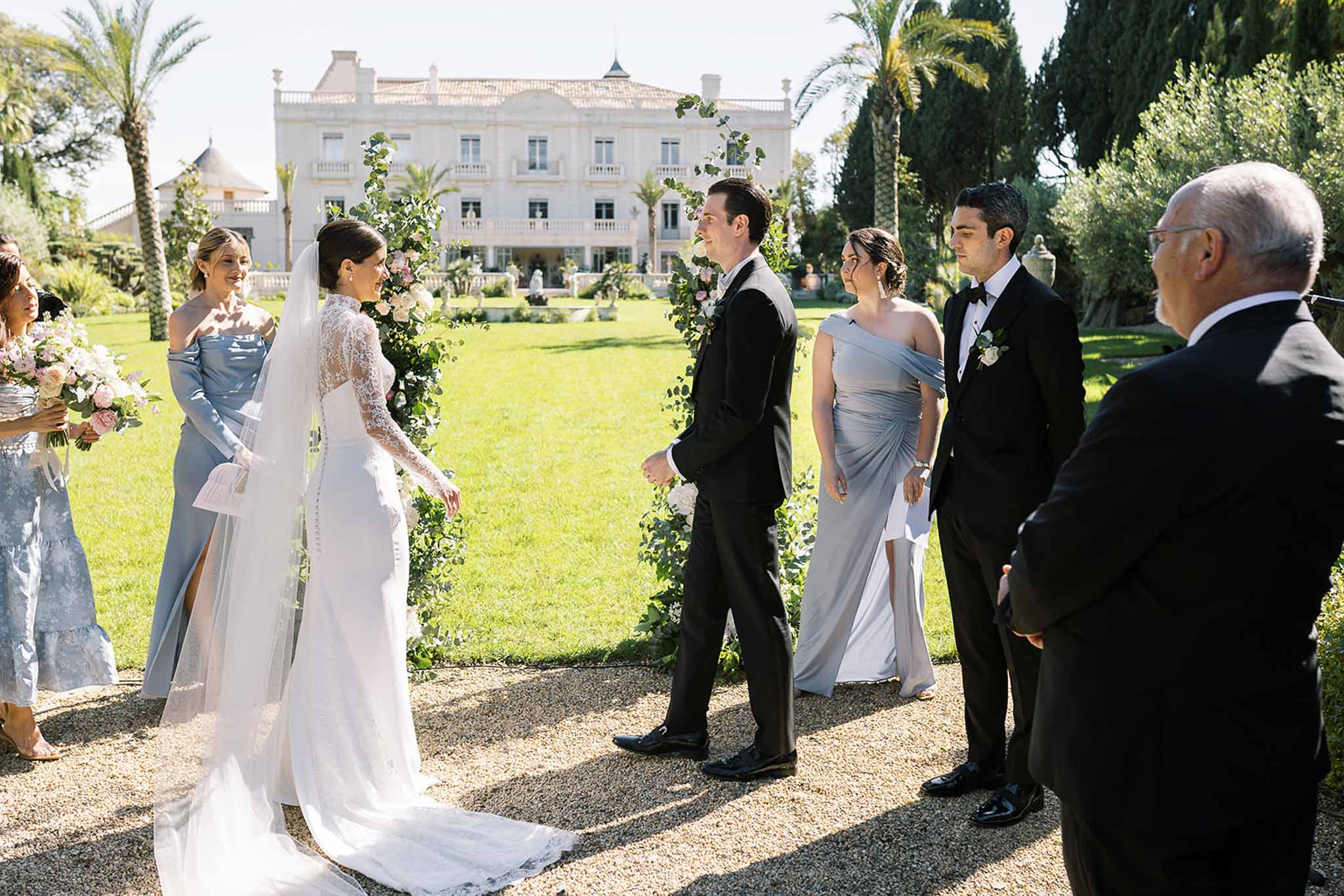 Wedding party at floral arch ceremony in front of white Belle Epoque villa with bridesmaids in blue gowns