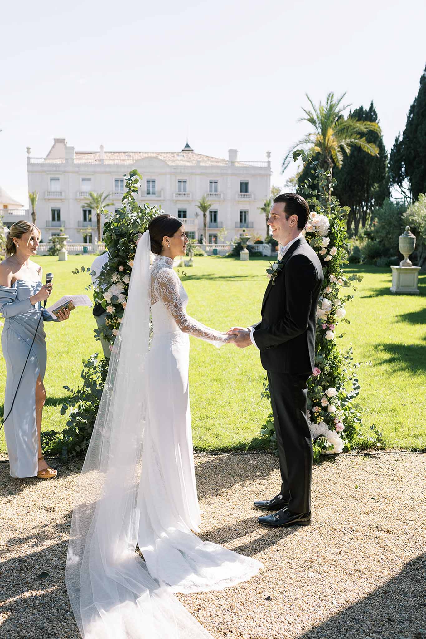 Bride and groom holding hands before circular floral arch during outdoor ceremony at neoclassical white villa