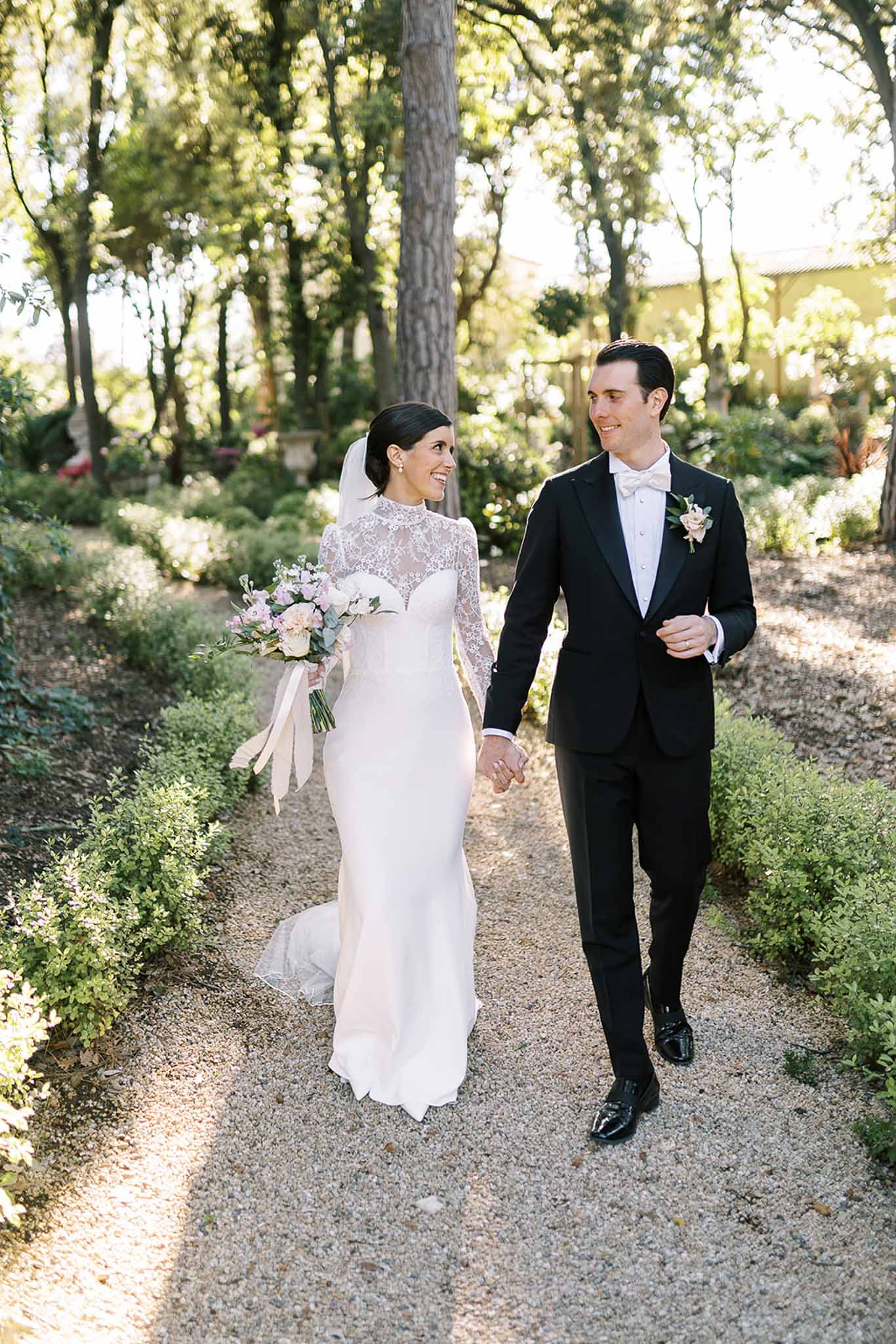 Bride and groom walking hand-in-hand on a gravel garden path lined with trimmed hedges