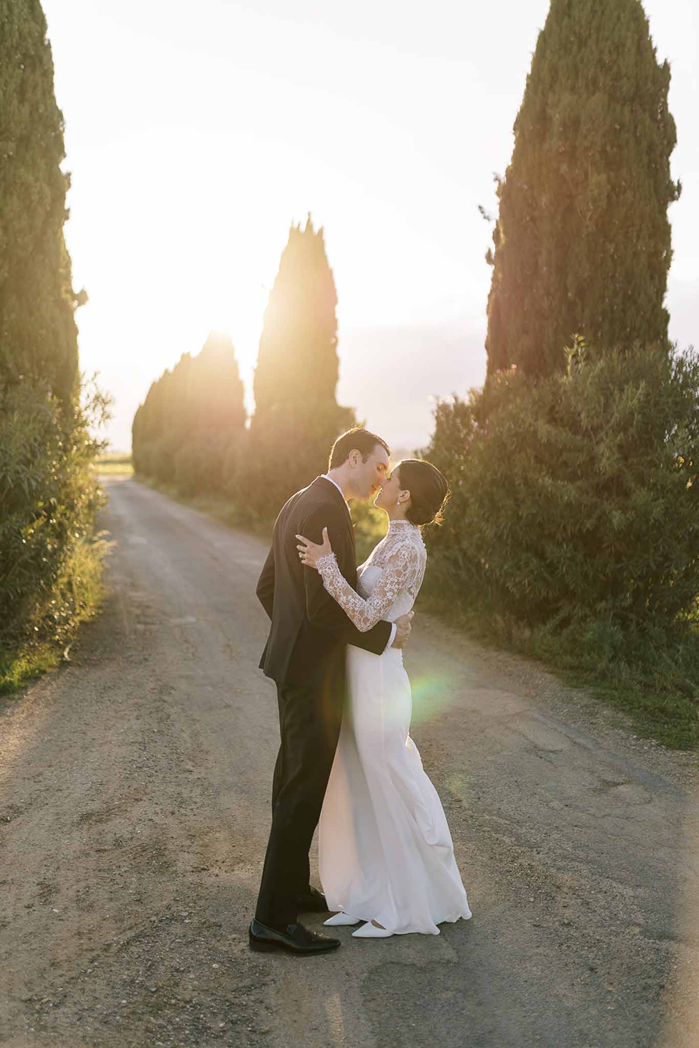 Couple kissing on cypress-lined gravel driveway with golden backlight and warm lens flare at sunset